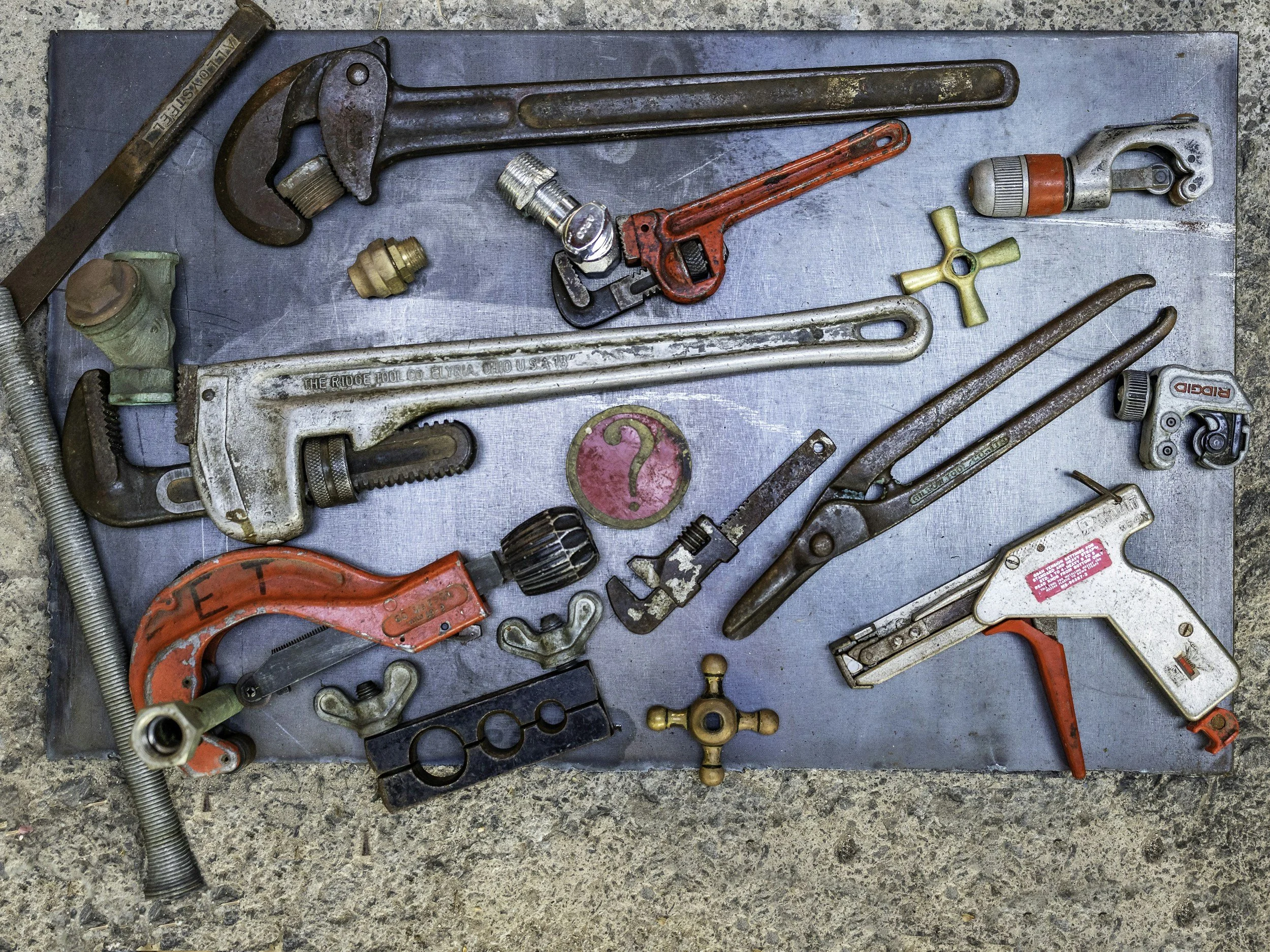 A metal surface covered with various old tools, including a pipe wrench, hacksaw, adjustable wrenches, pipe fittings, a saw blade, and other hardware, all arranged in a scattered manner on a concrete floor.