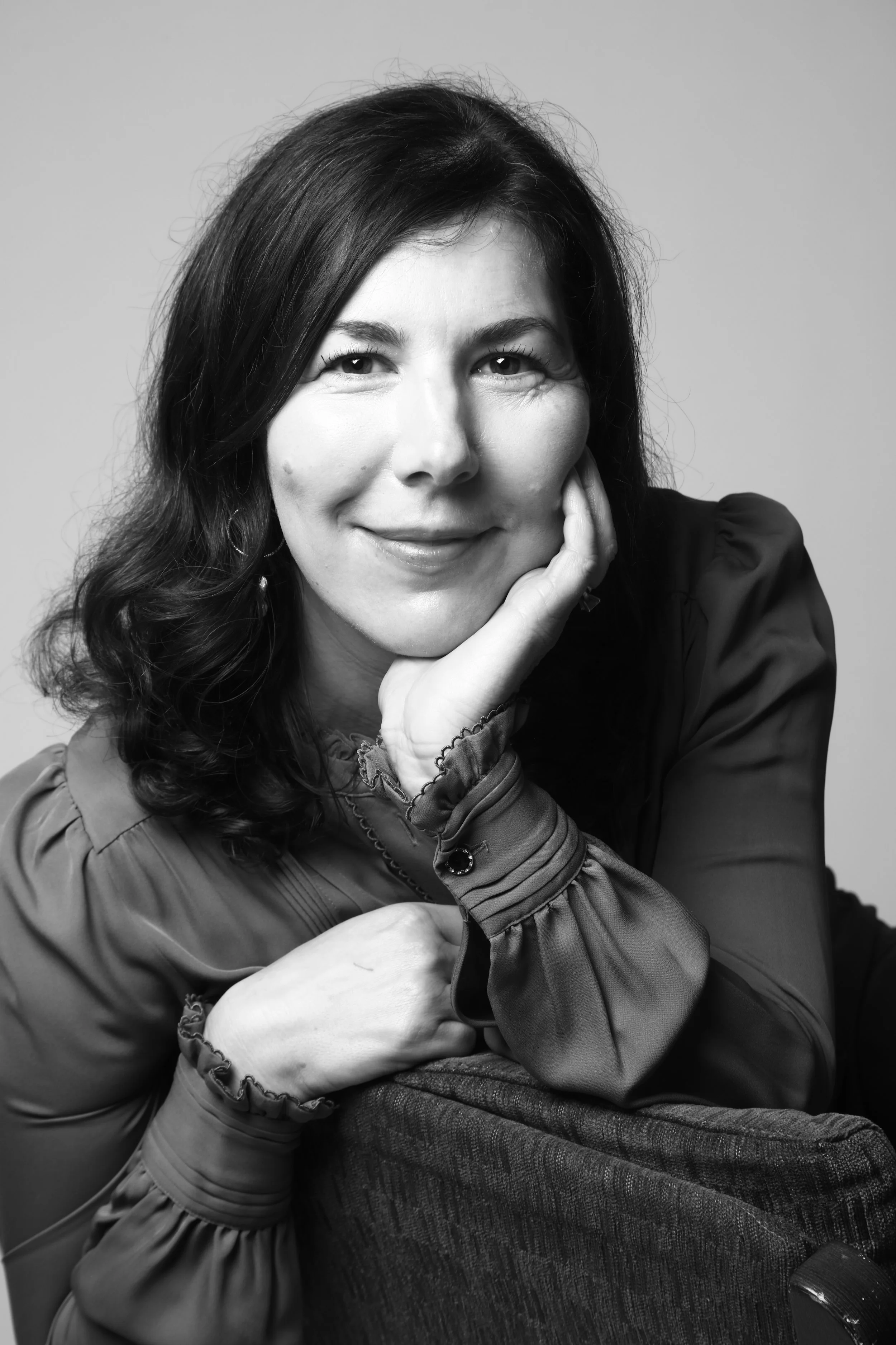 Black and white close-up photograph of a woman with dark hair, smiling gently, resting her chin on her hand, wearing a long-sleeved, ruffled blouse with a button on the cuff, sitting with her arm draped over a textured armrest.