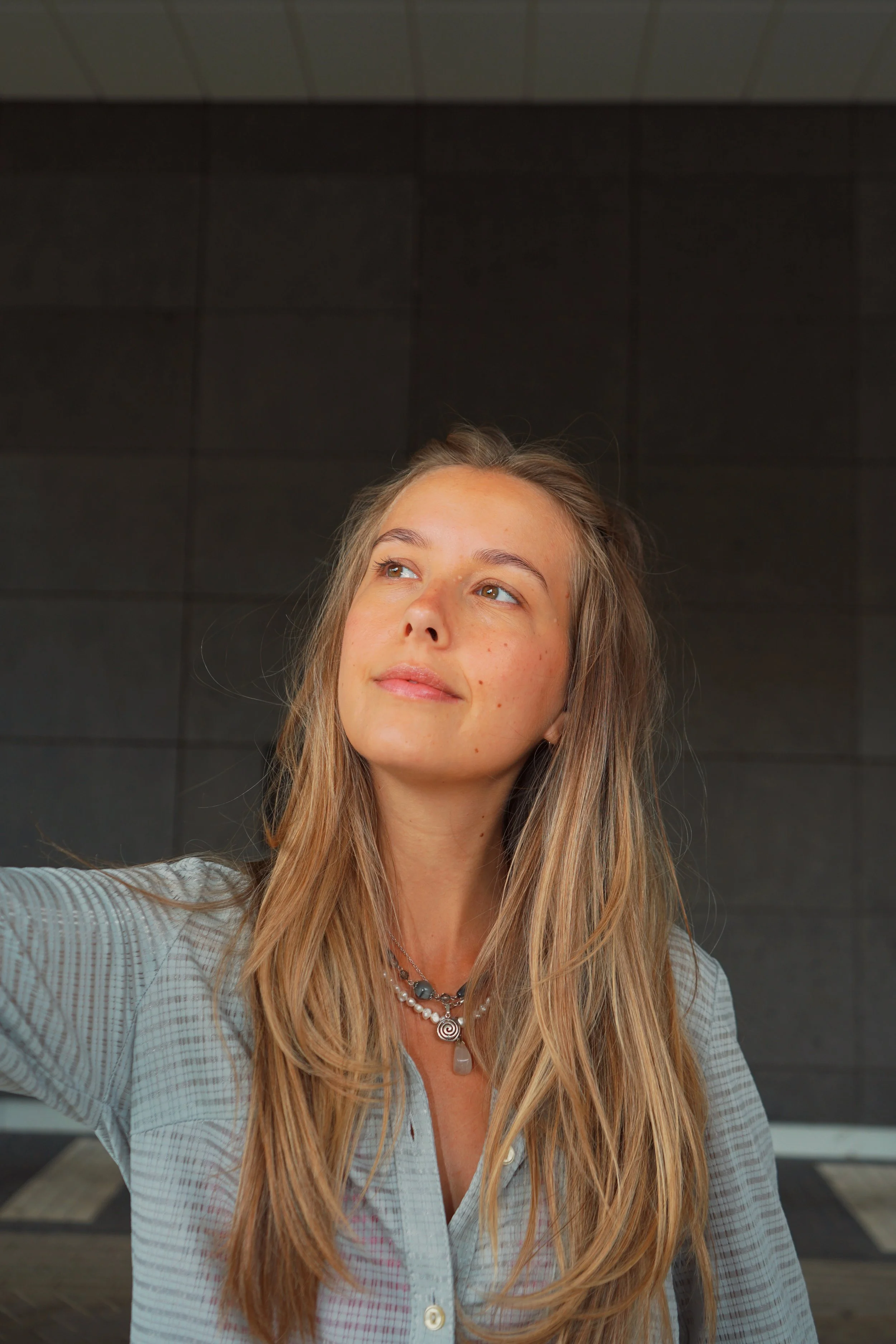 A woman with long blonde hair taking a selfie indoors, standing in front of a black tiled wall, wearing a light blue checkered shirt and layered necklaces.