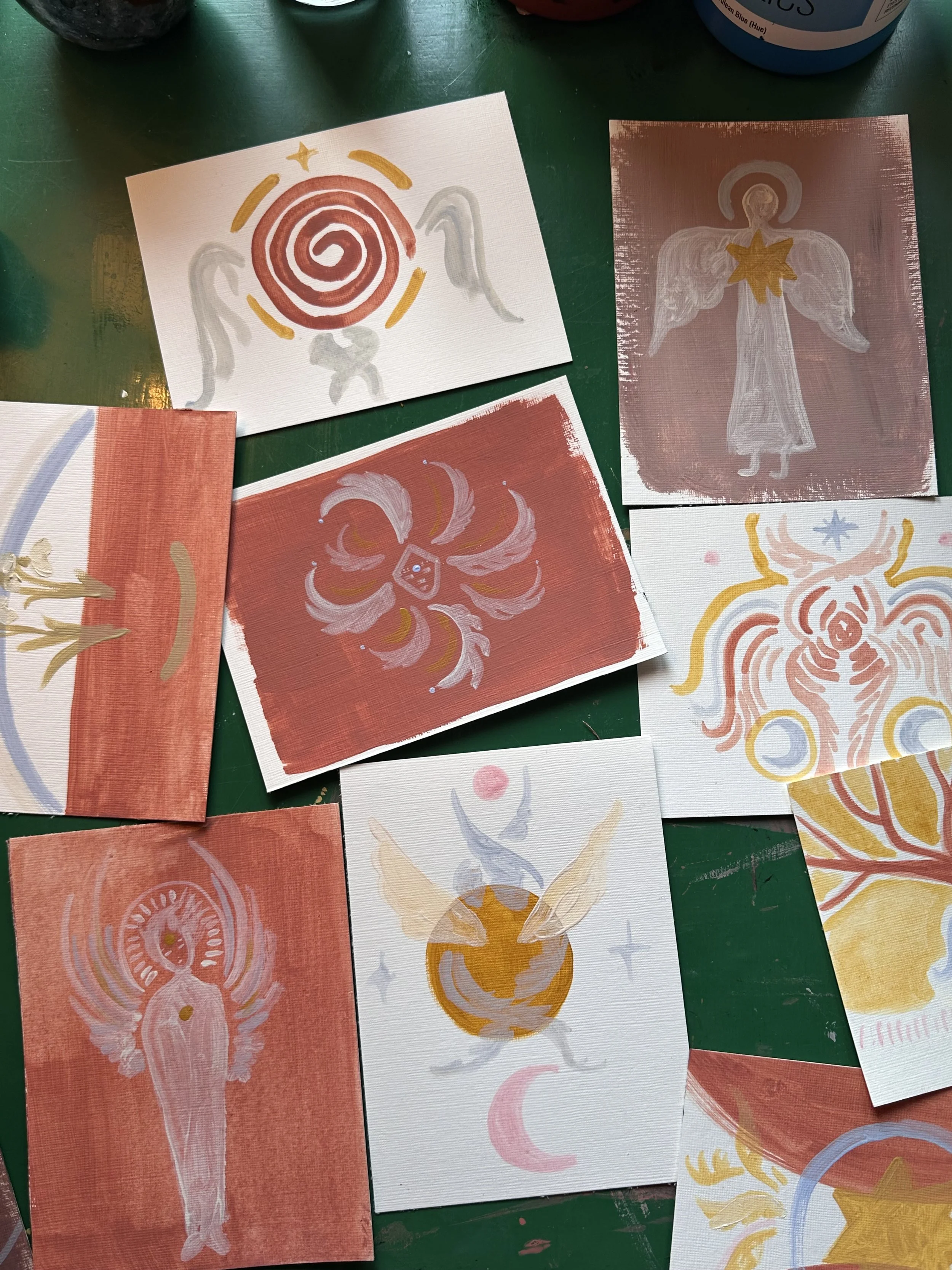 Multiple abstract paintings with angelic and celestial themes, featuring wings, stars, and swirling patterns on a green surface.