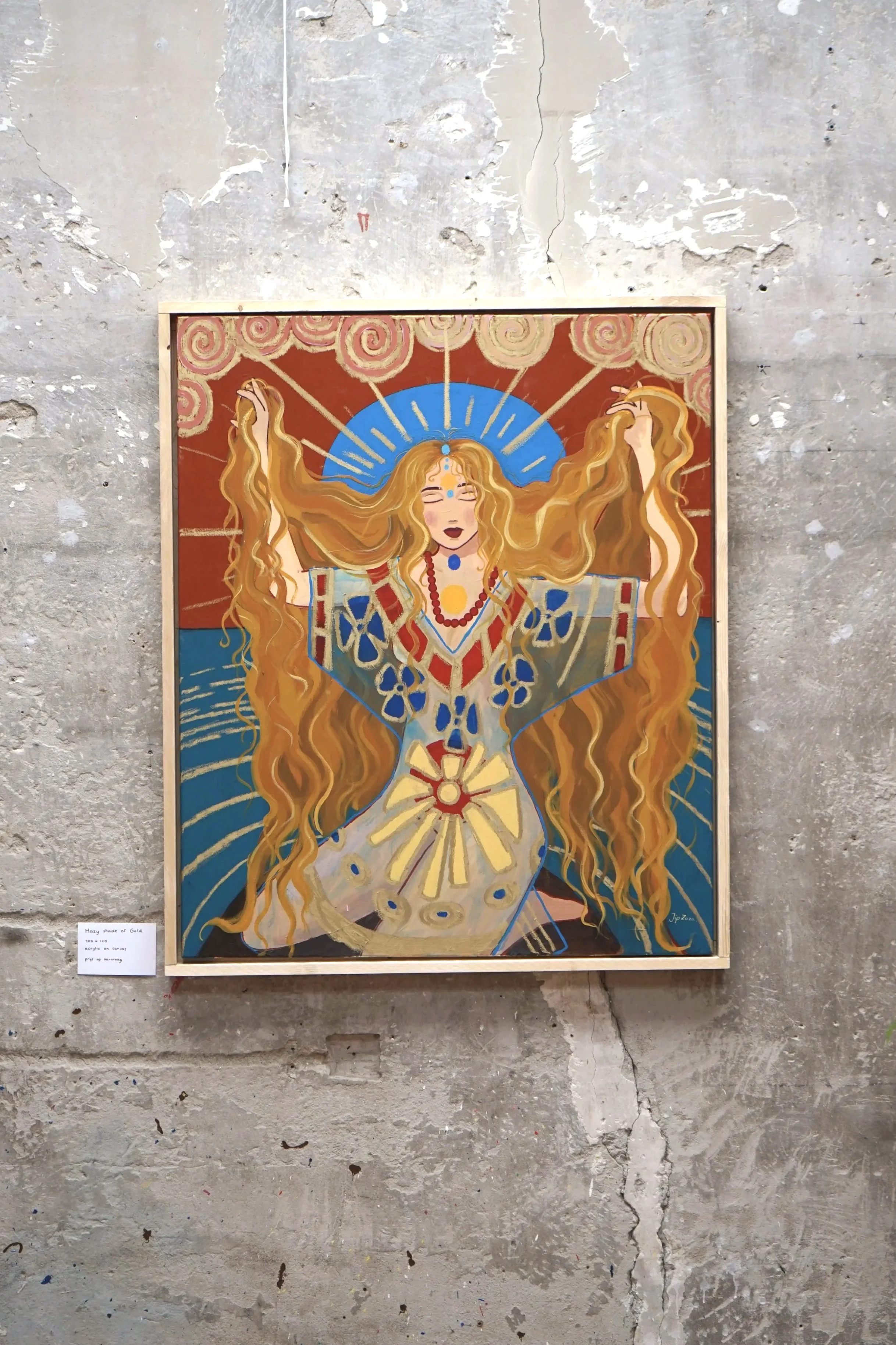 A colorful painting of a woman with long, flowing golden hair, closed eyes, and a serene expression. She has various decorative elements around her, including a blue halo with radiating lines, a red necklace, and intricate blue and red patterns on her clothing. The background features stylized cloud shapes and a sunset-like sky. The painting is displayed on a rough, gray, textured wall.