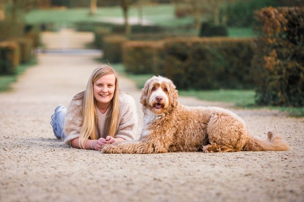 A woman with long blond hair lying on her stomach on a gravel path, smiling, next to a large cobberdog lying down, in a park setting with trees and bushes in the background.