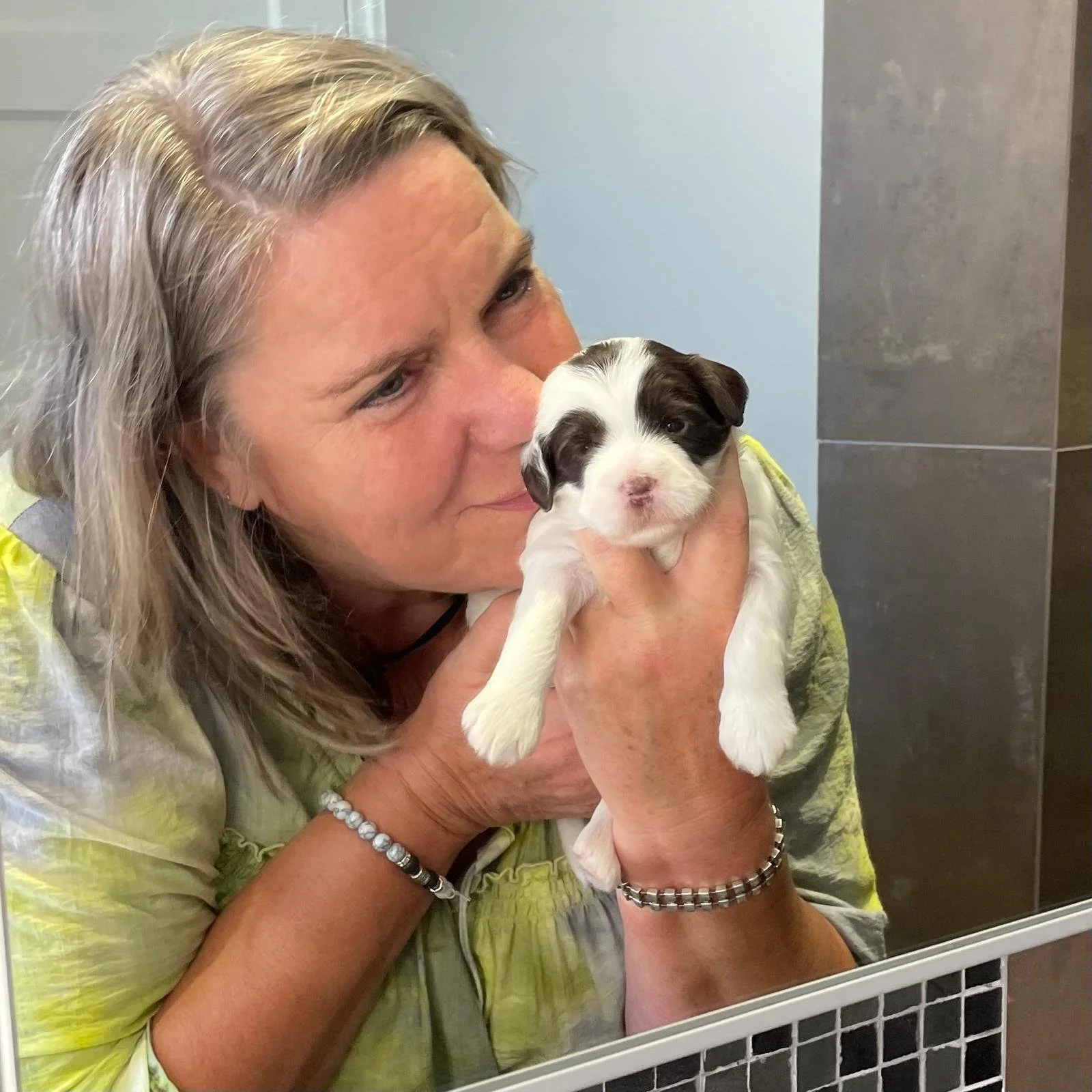 A cobberdog breeder with blonde hair holding a small black and white cobberdog puppy, smiling at the cobberdog puppy, in a room with gray tile and a mirror.