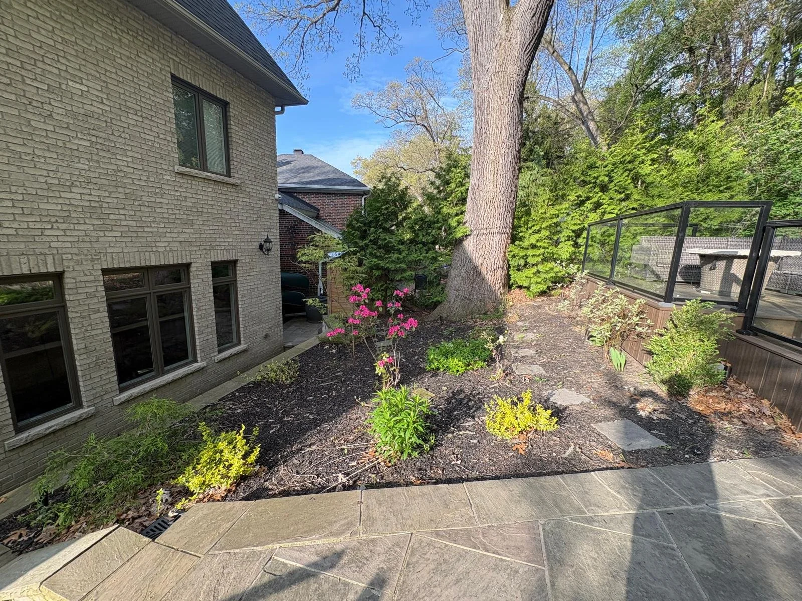 Backyard garden with a stone patio, flower bed with pink, yellow, and green plants, large tree, and a screened-in porch on the right, adjacent to the brick house with multiple windows.