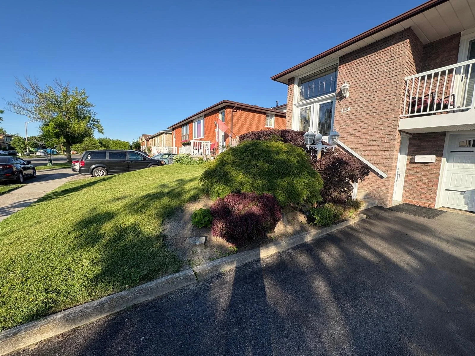 Residential neighborhood with brick townhouses, parked cars, green lawn, and well-maintained bushes under a clear blue sky.