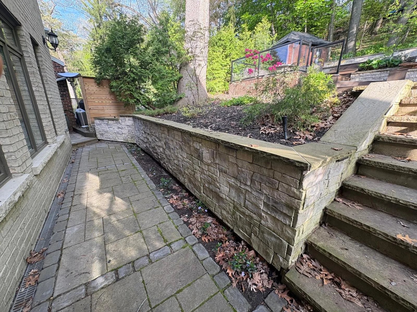 A backyard patio with a paved walkway, a stone retaining wall, and wooden stairs leading up to a garden area with trees and plants. There is a house to the left with windows and outdoor lighting.
