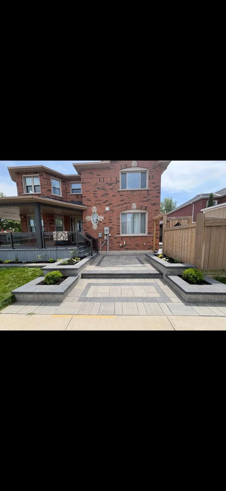 Backyard patio with a brick house in the background, featuring windows and a small porch with a protective railing, surrounded by flower beds and a wooden fence.