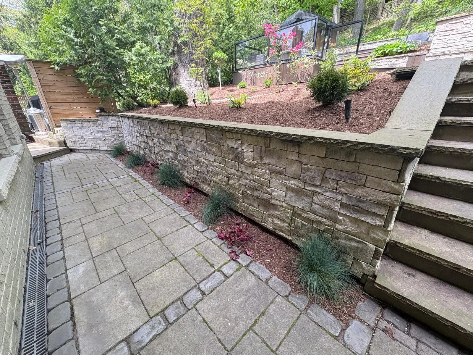 A backyard with a paved patio area, a stone retaining wall, and a raised garden bed with small bushes and pink flowers. Wooden stairs lead up to the garden. Green trees and foliage are in the background.