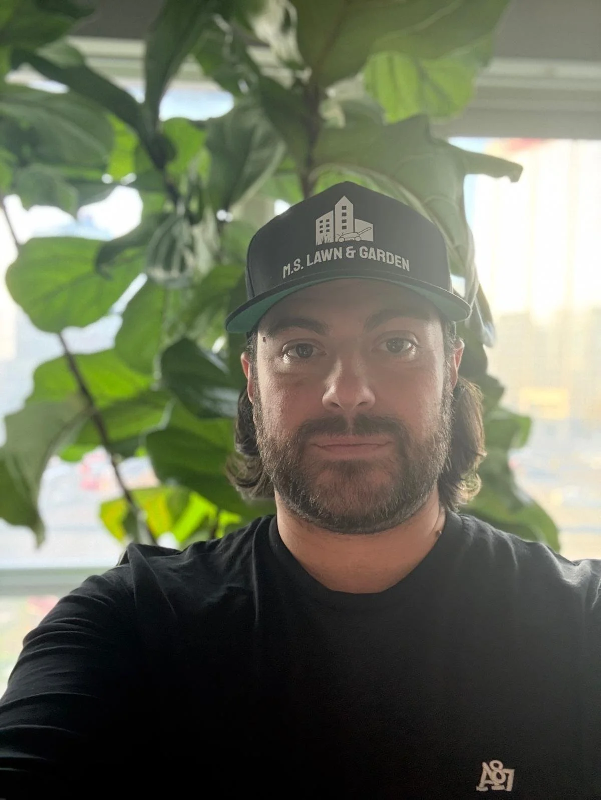 A man with dark hair and a beard wearing a black cap that says "M.S. LAWN & GARDEN" and a black t-shirt, standing in front of large green leaves indoors.