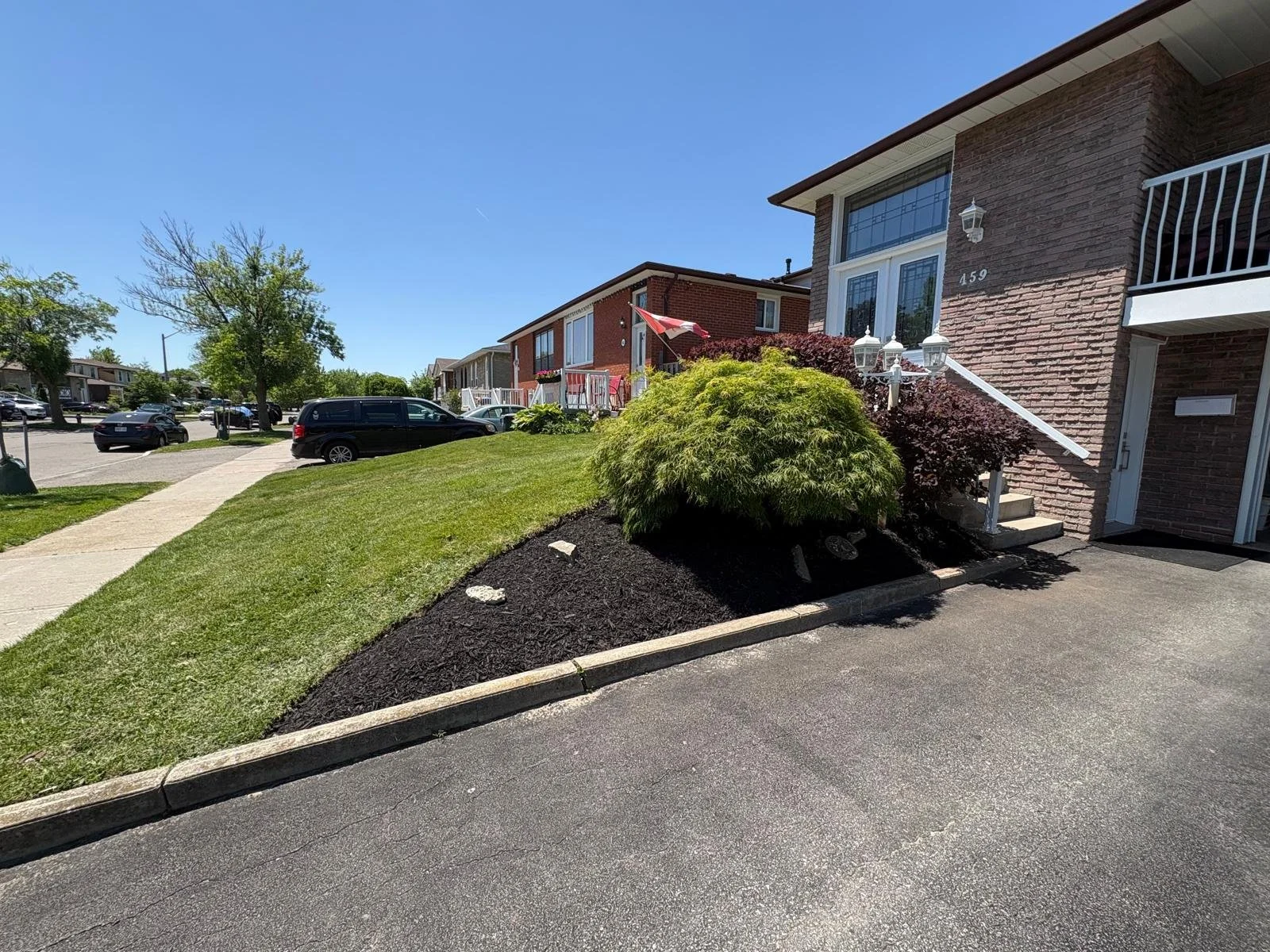 Residential apartment building with landscaped front yard, sidewalk, parked cars, and a blue sky.