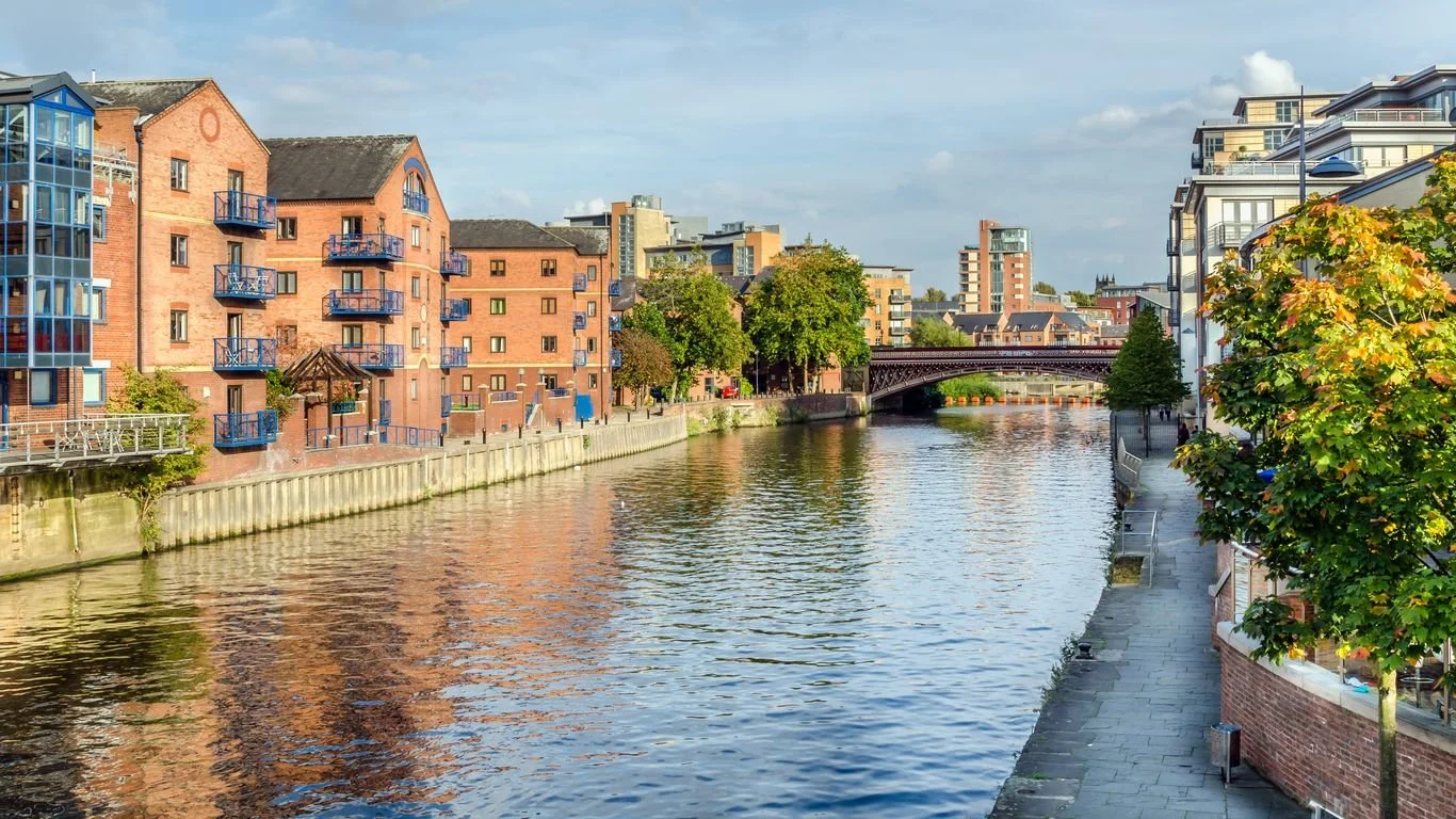 Cityscape with a canal, brick buildings with blue balconies on the left, modern white buildings on the right, a bridge in the distance, and trees along the walkway.