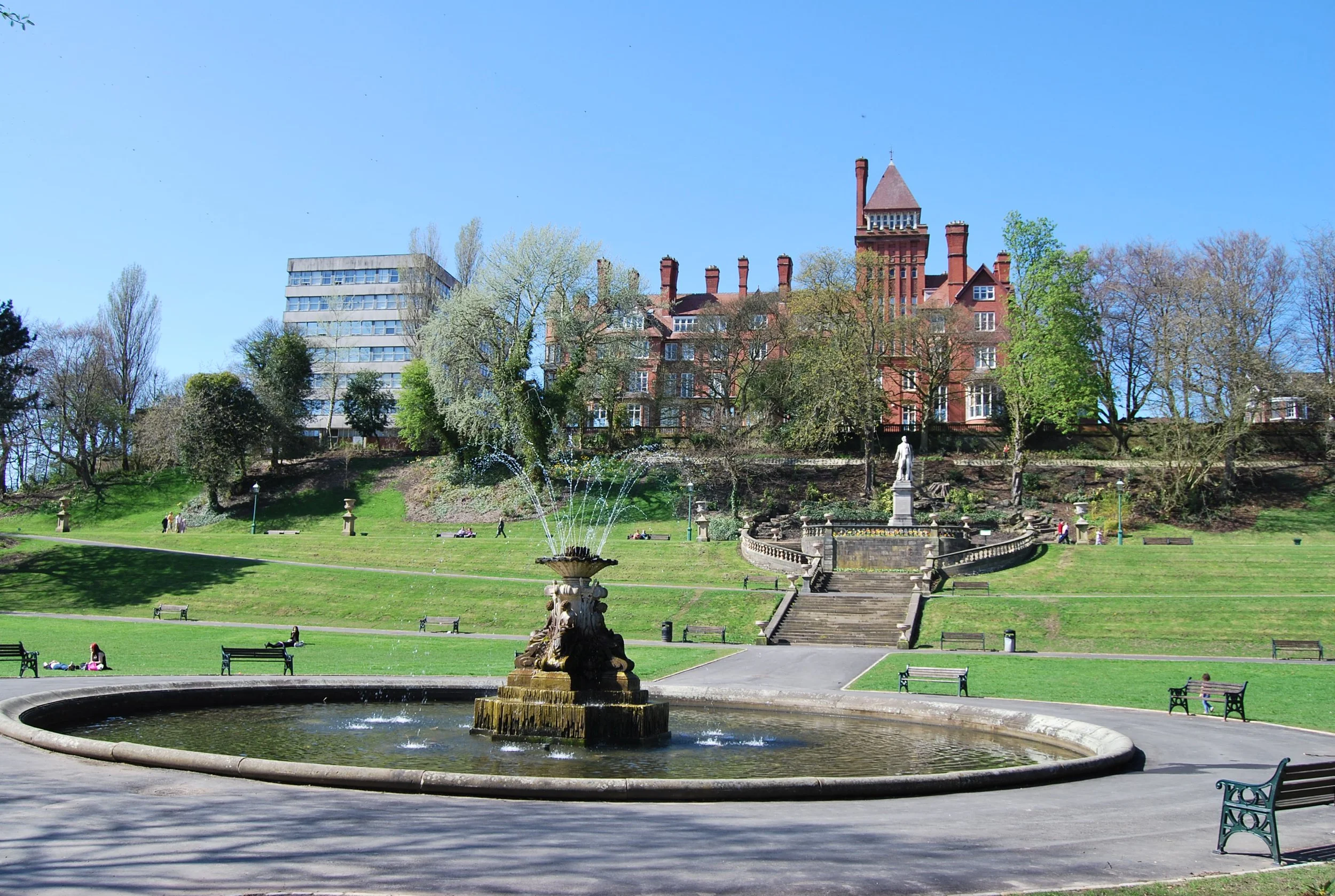 A park with a fountain in the foreground, trees and a grassy area with benches, and a large historic red brick building with a tower in the background against a clear blue sky.