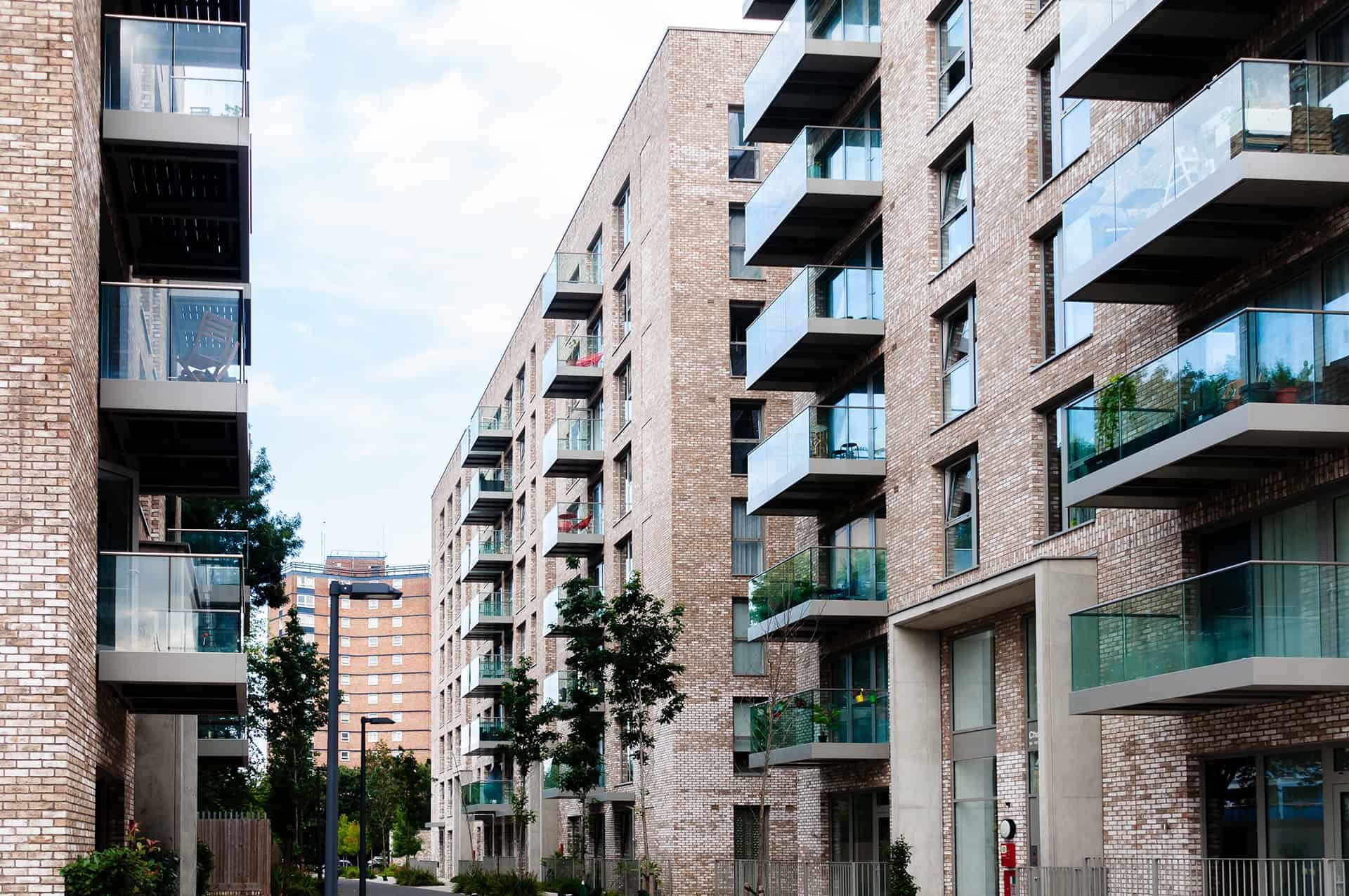 Modern multi-story apartment buildings with brick facades and glass balconies, trees and streetlights in an urban residential area.