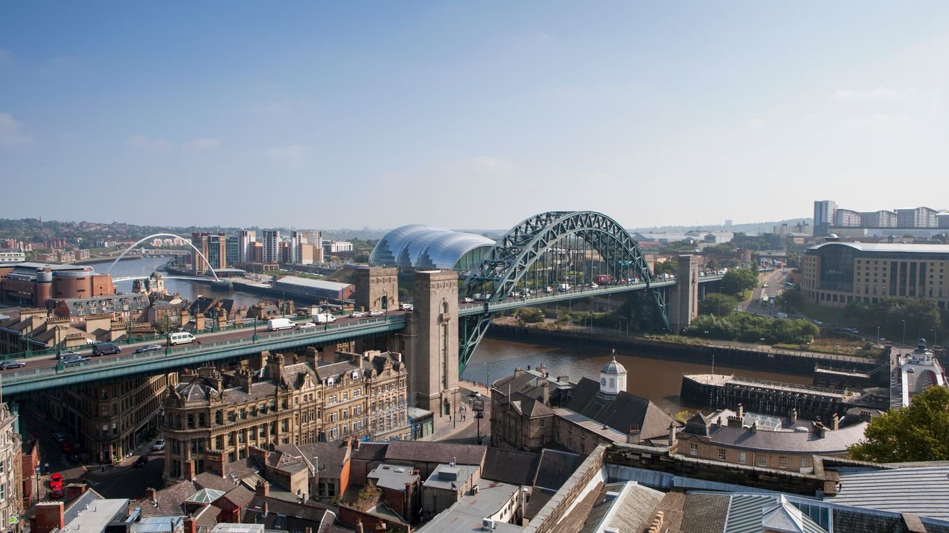 A cityscape view featuring a bridge over a river, with multiple buildings and a modern structure with a curved roof in the background.