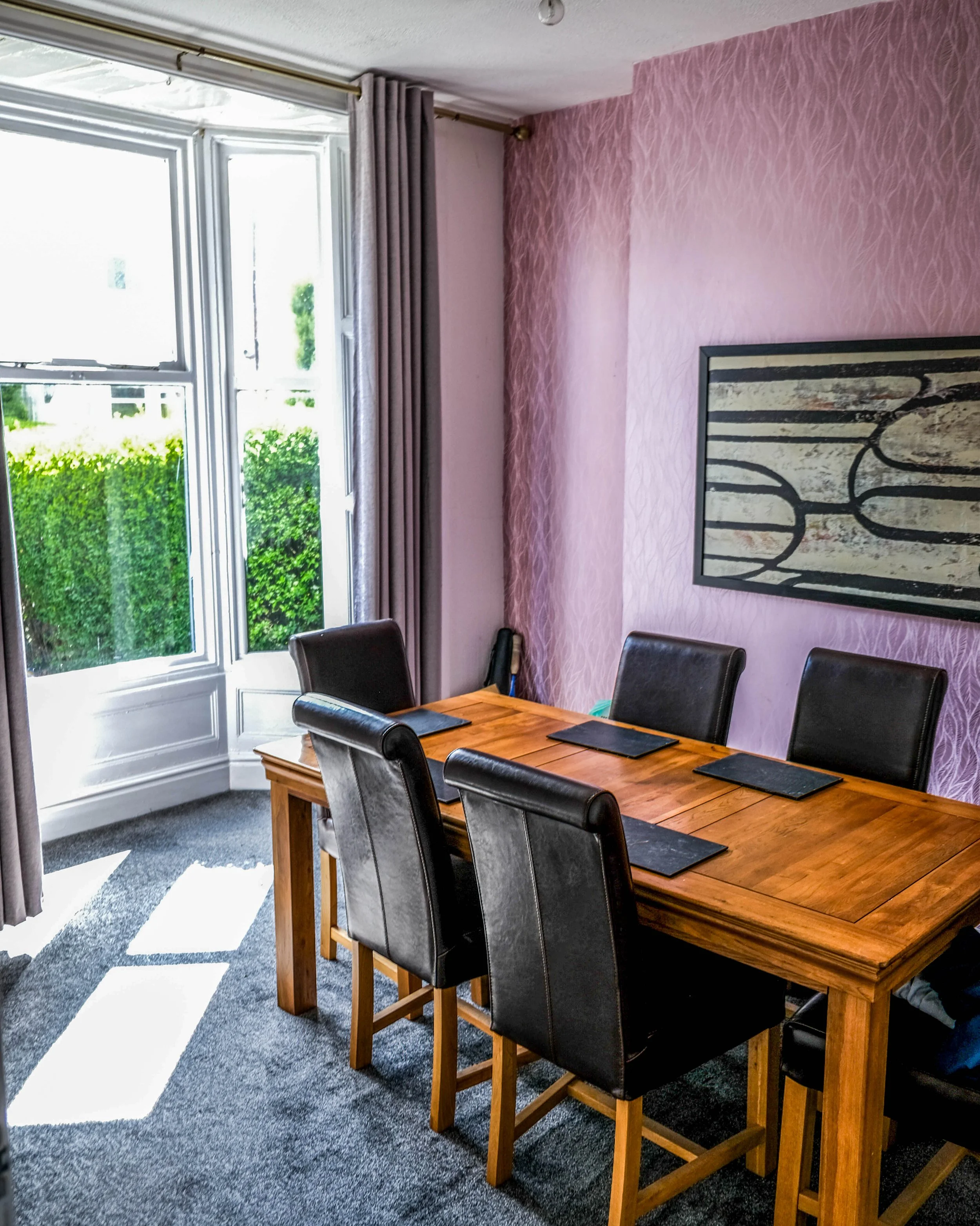 A dining room with a wooden table and six black leather chairs. Large windows with gray curtains, pink textured walls, and a piece of abstract wall art.