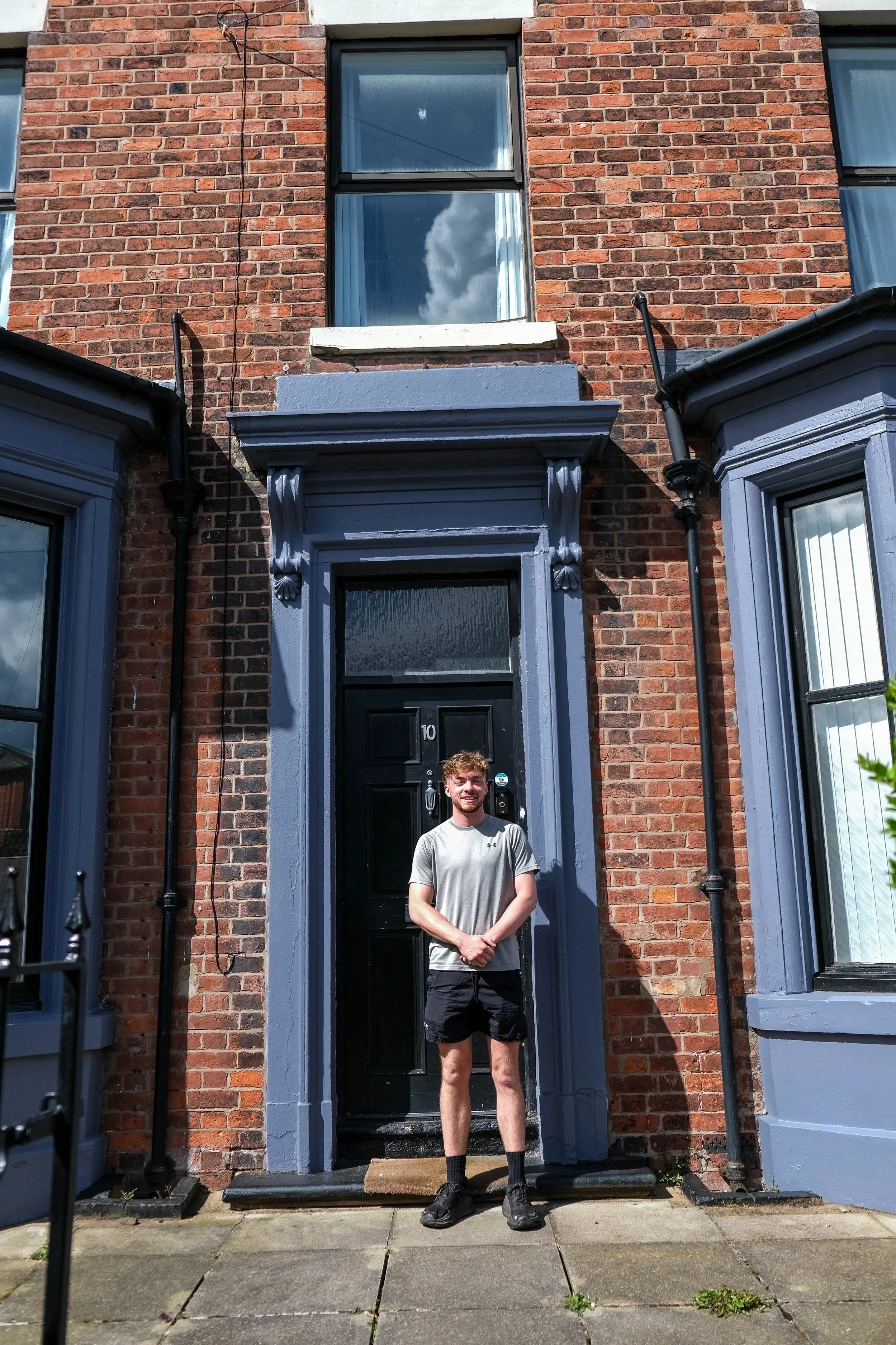 Young man standing on front porch of brick townhouse, smiling, wearing a gray t-shirt, black shorts, and black shoes, with front door numbered 10, flanked by two windows with blue trim and white curtains.