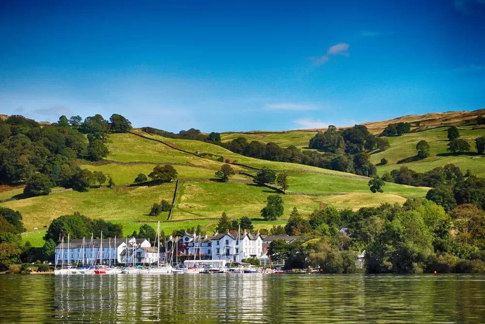 Scenic view of a lakeside town with sailboats docked, surrounded by green rolling hills under a blue sky.