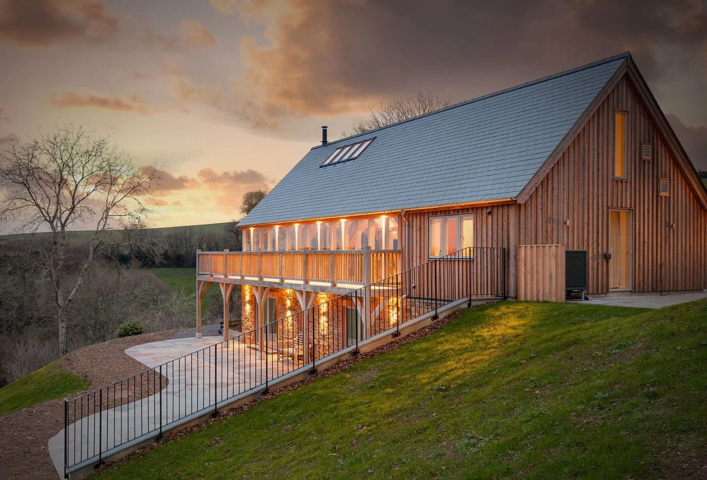 A modern wooden house on a hillside at sunset, with large windows and a balcony illuminated from inside, overlooking a landscape with trees and hills.