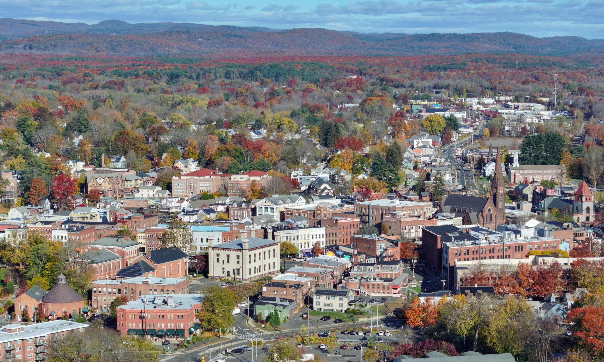 An aerial view of a small town with colorful fall trees, old and modern buildings, church steeples, and rolling hills in the background under a partly cloudy sky.