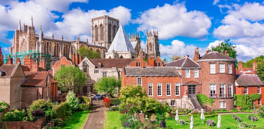 A view of a historic cathedral in the background with gothic architecture, surrounded by traditional brick houses and a lush green garden with outdoor seating in the foreground.