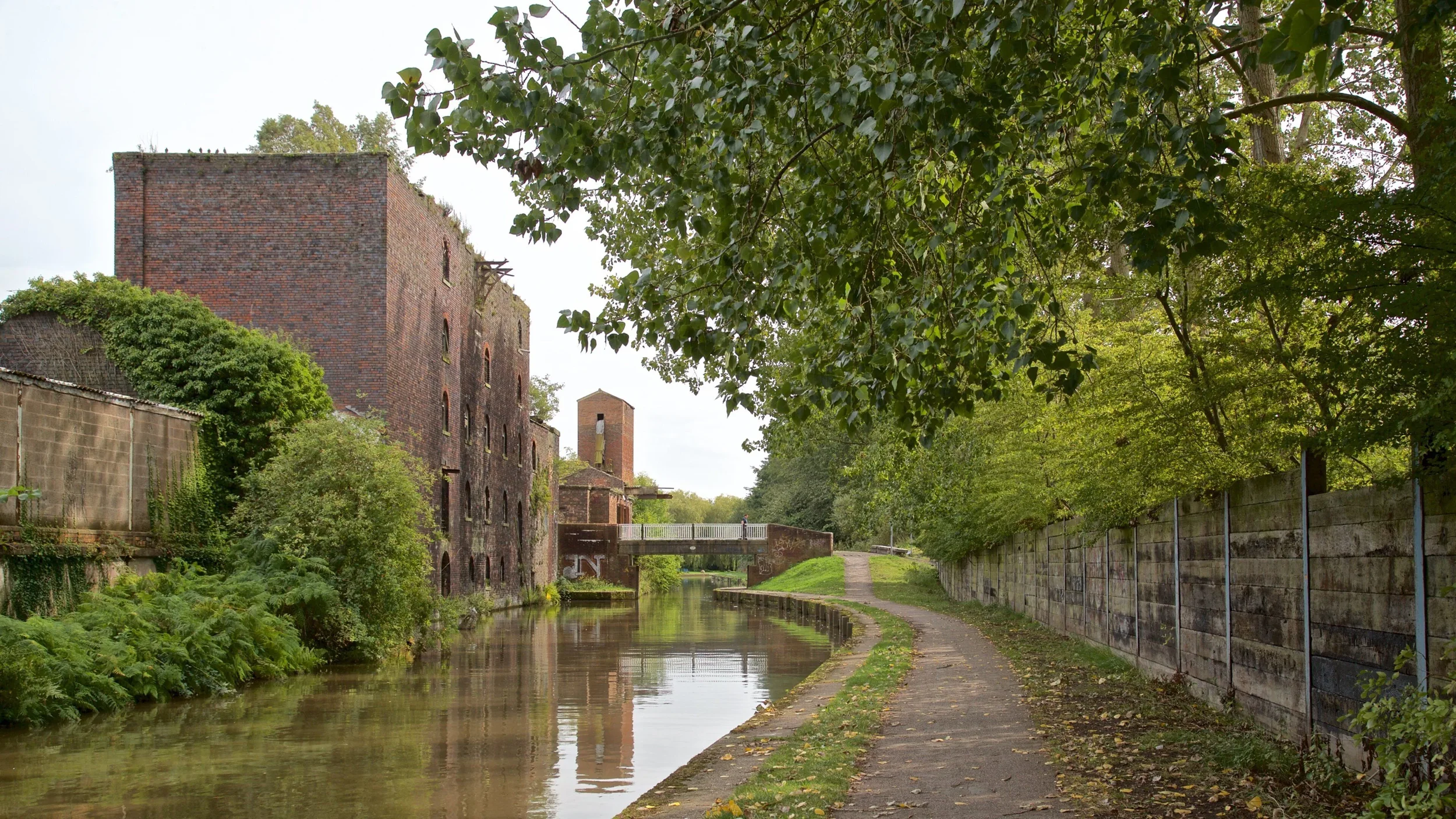 A brick building next to a canal with a walking path, surrounded by green trees and foliage.