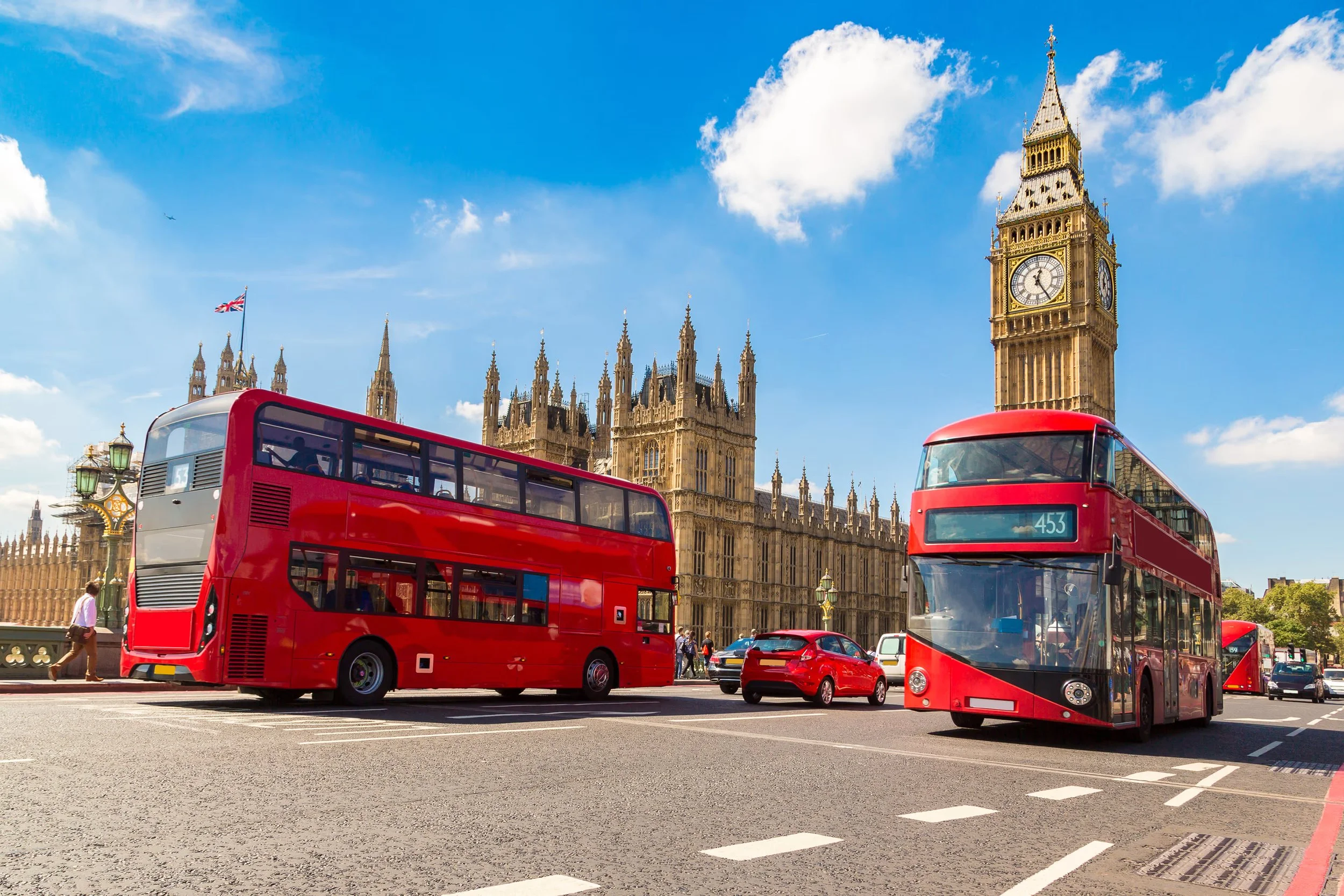 Two red double-decker buses on a street with the Houses of Parliament and Big Ben in the background under a blue sky with some clouds.