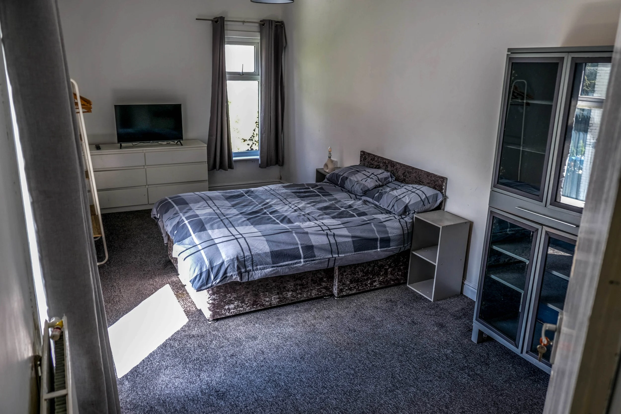 A bedroom with a bed covered in a gray and black checkered blanket, two pillows, a nightstand with a lamp, a sitting cabinet with glass doors, a small empty shelf, a dresser with a TV on top, a window with gray curtains, and a coat rack visible on the left side.