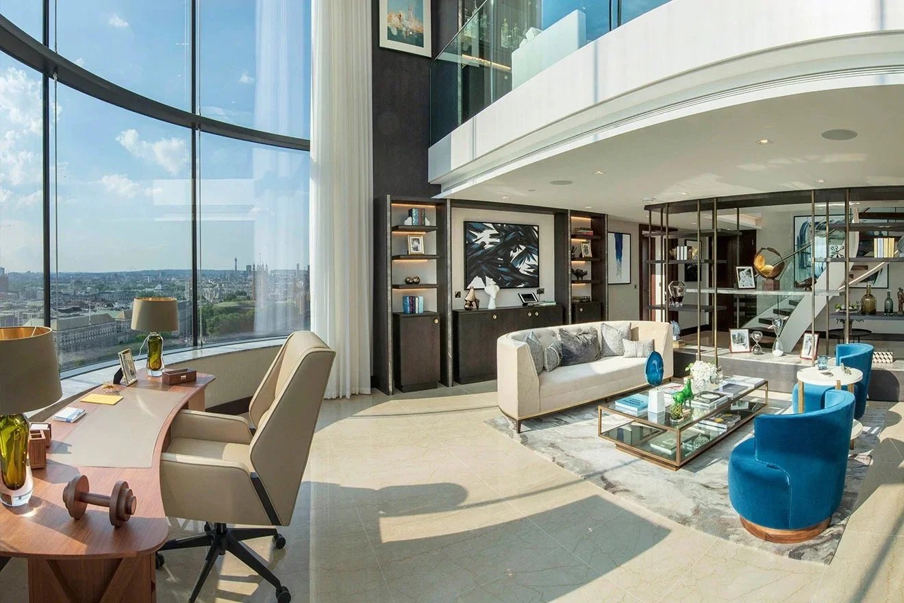 Modern high-rise living room with large curved window, beige desk and chairs, white sofa, blue armchairs, glass coffee table, black bookshelves, artwork, and city view.