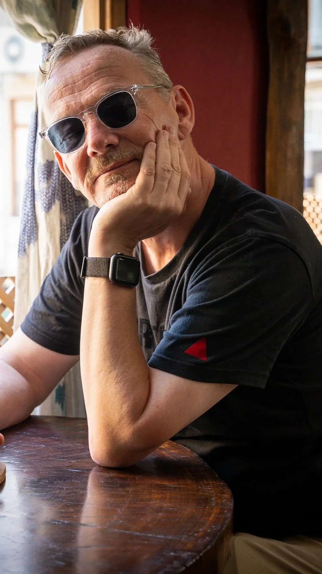 A man with gray hair, wearing sunglasses, a black t-shirt, and a smartwatch, sitting at a wooden table with a contemplative expression, resting his face on his hand. Istanbul, Turkey
