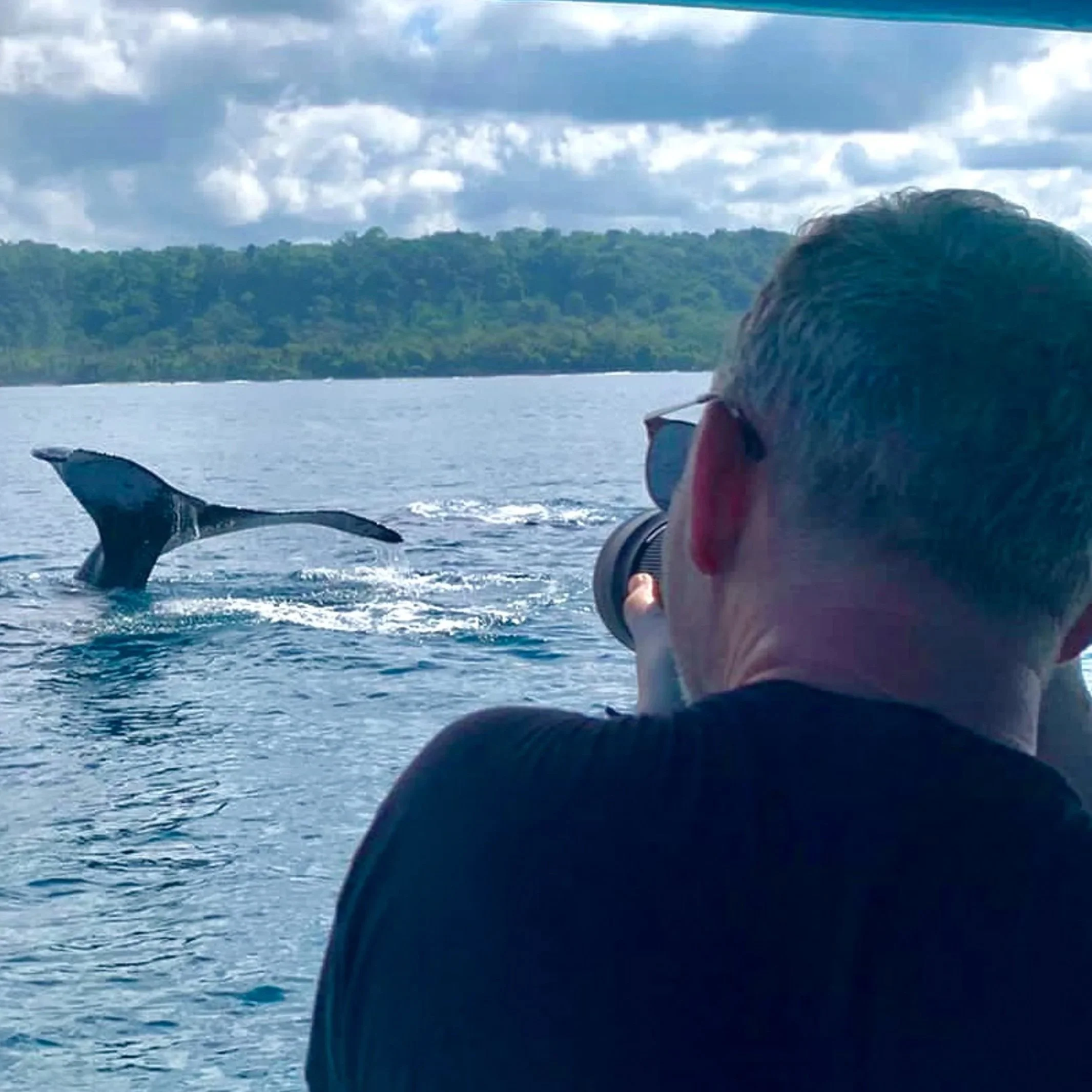 A person taking a photo of a humpback whale tail fin in the Osa Peninsula, Costa Rica, tree-covered shoreline and cloudy sky in the background.