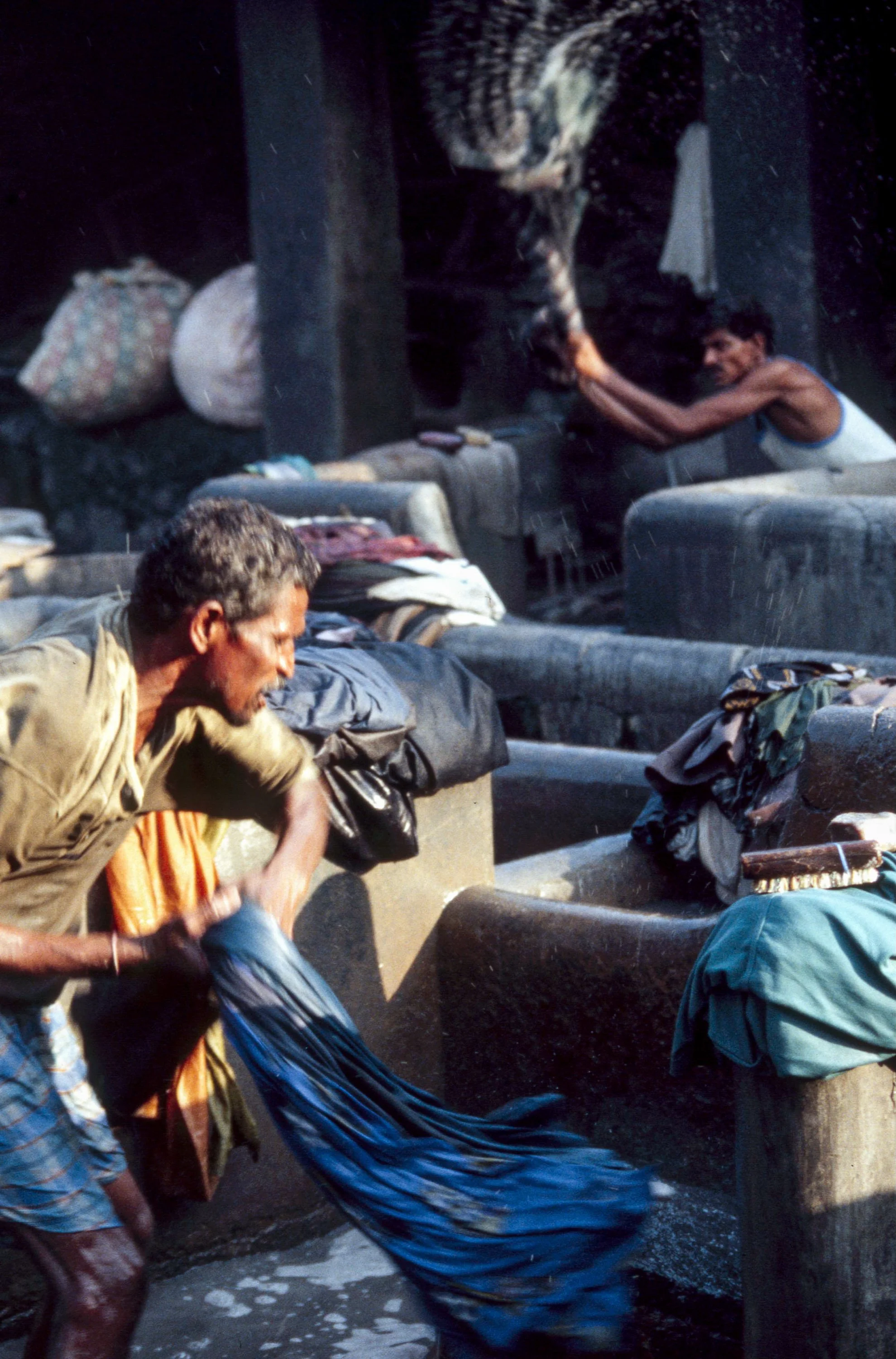 People washing clothes in an outdoor water basin, some wearing traditional clothing, in a lively scene.