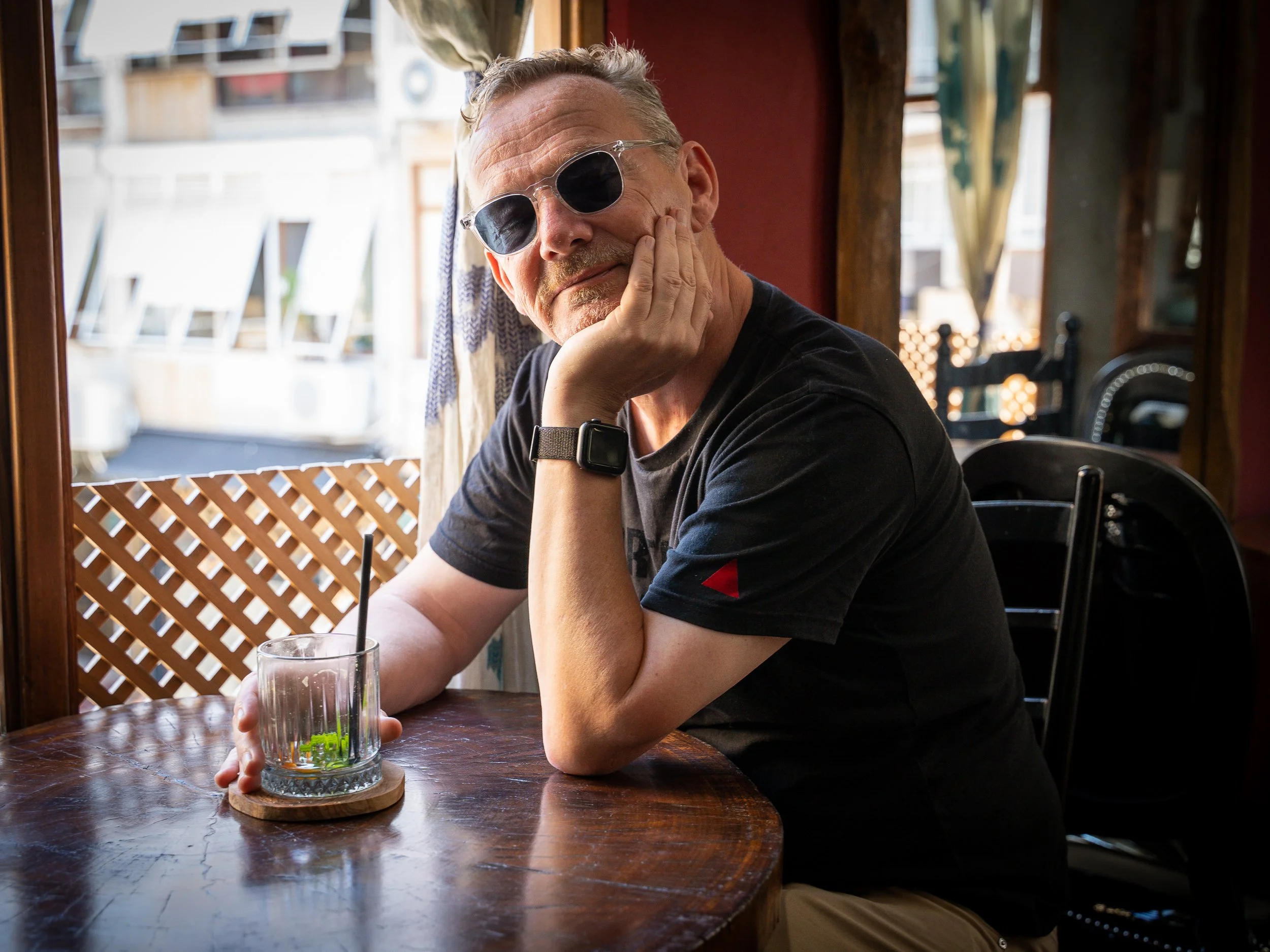 A man with sunglasses and a watch sitting at a wooden table in a casual restaurant, resting his face on his hand, with a glass of ice water with lime and a straw in front of him. Istanbul, Turkey