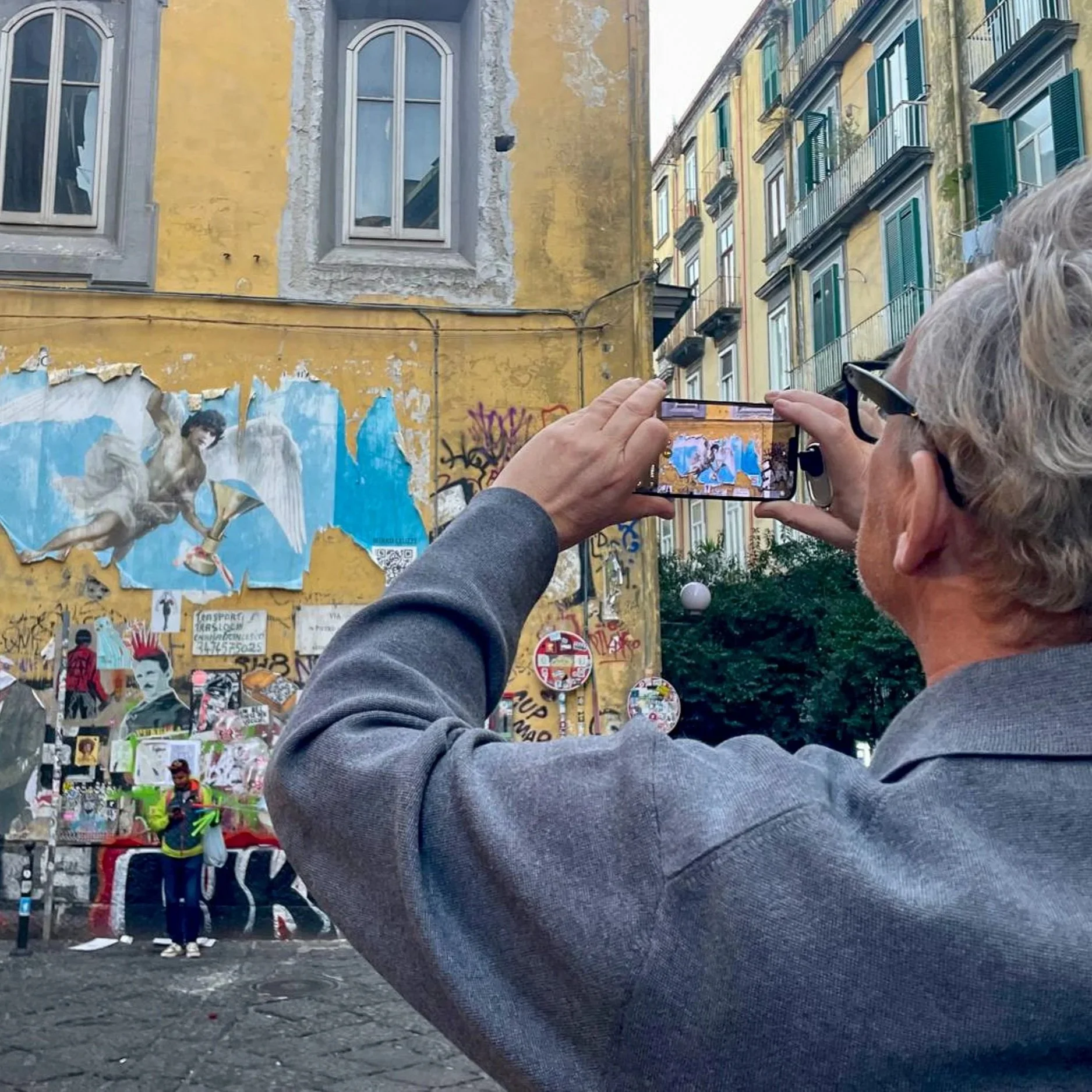 Person taking a photo of a colorful street mural with a smartphone, in front of a yellow building with green window shutters, graffiti, posters, and some people in the background.