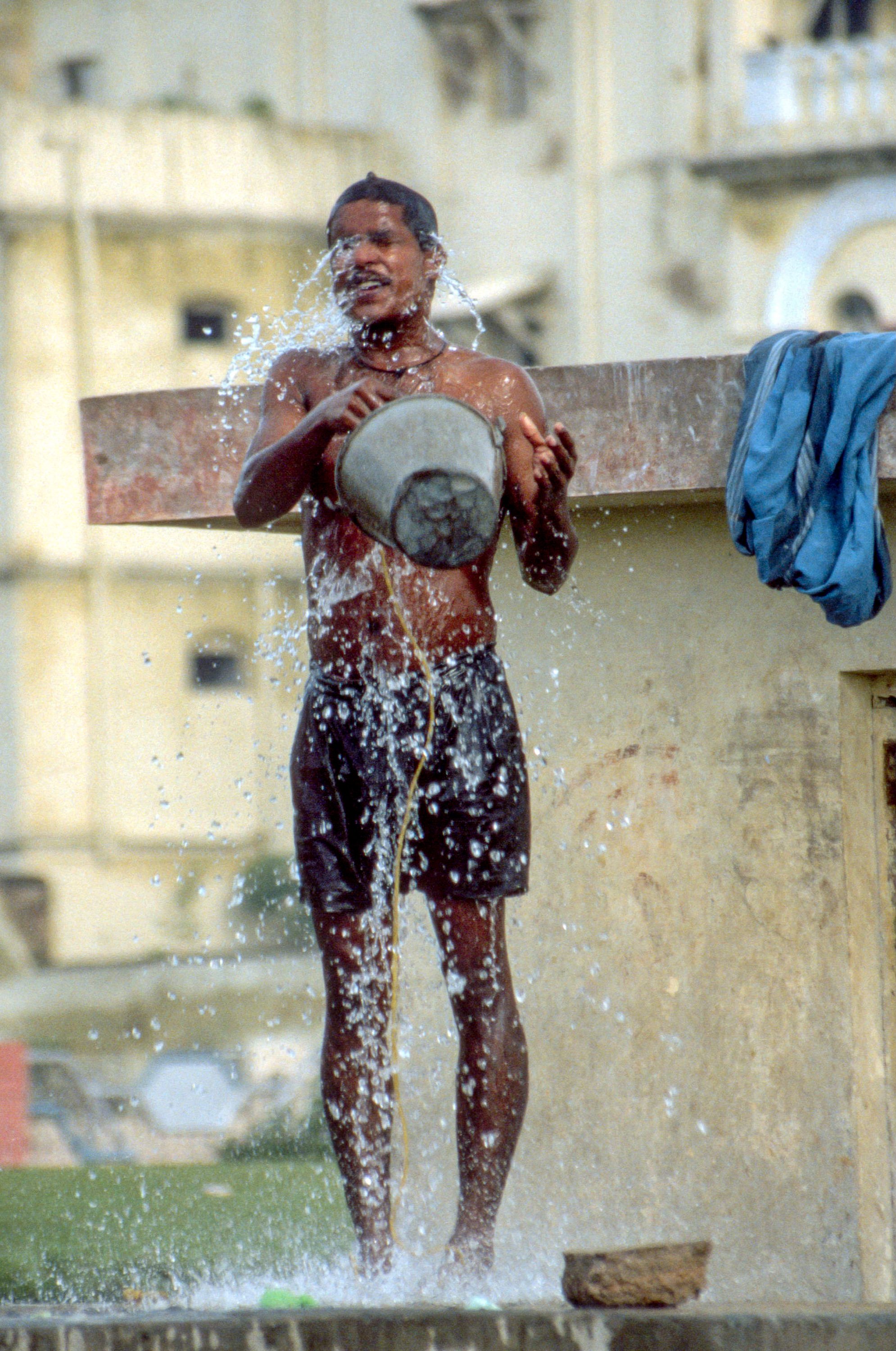 A young boy taking a shower outdoors while holding a bucket and smiling in Chennai India.
