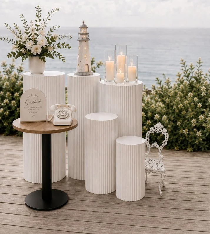 Decorative outdoor setup with white textured pedestals, a vase of white flowers, a miniature lighthouse, lit candles in glass holders, a vintage typewriter, a sign for guestbook, a small round table, and an ornate white chair against a backdrop of the sea and sky.