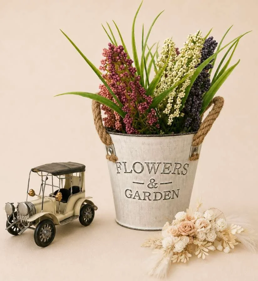 A decorative flower arrangement in a white bucket labeled "Flowers & Garden," with a small vintage toy car next to it and a cream-colored flower bouquet in front.