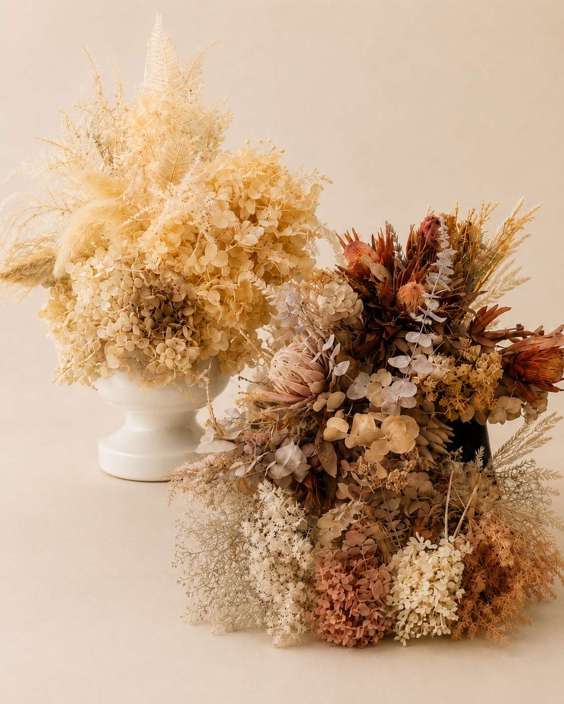 Two vases filled with dried beige, brown, and cream-colored flowers and foliage, arranged on a neutral background.