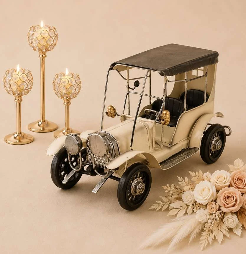 A vintage toy car model with a white body and black roof, three tall gold candles, and a bouquet of pink and white roses with beige leaves on a neutral background.