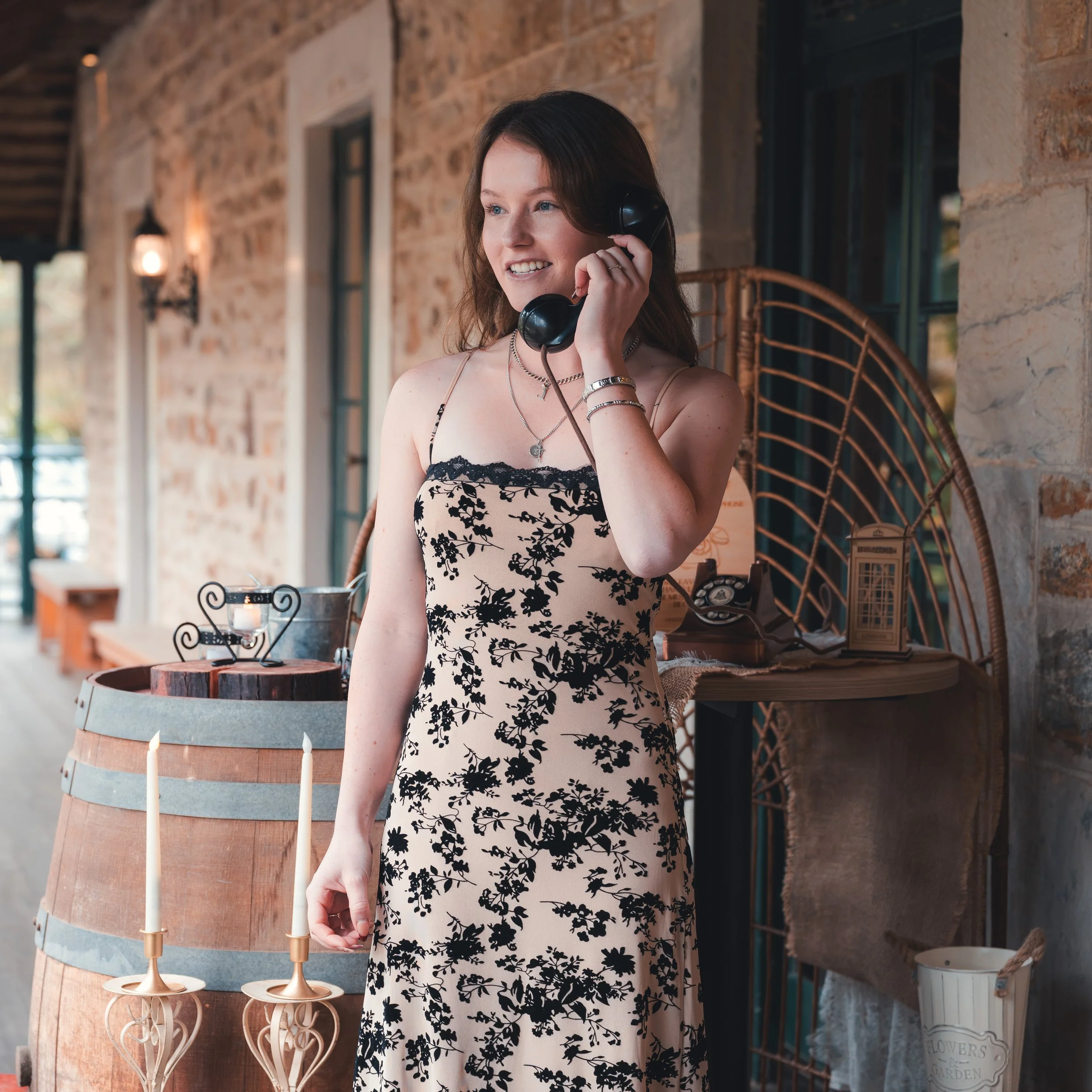 A smiling woman with brown hair, wearing a black and white floral dress, holding a vintage black telephone receiver to her ear inside a rustic brick-walled room with wooden accents.