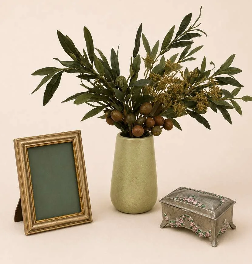 A beige surface with a small gold-framed photo frame, a green vase with leafy branches and berries, and a small silver jewelry box with floral embroidery.