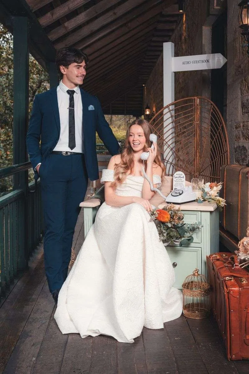 A young woman in a white dress is sitting on a desk using a vintage rotary phone, smiling and talking. A young man in a navy suit is standing beside her, smiling and touching her shoulder. The scene is set in a rustic indoor space with a stone wall and a sign pointing to an audio guestbook.