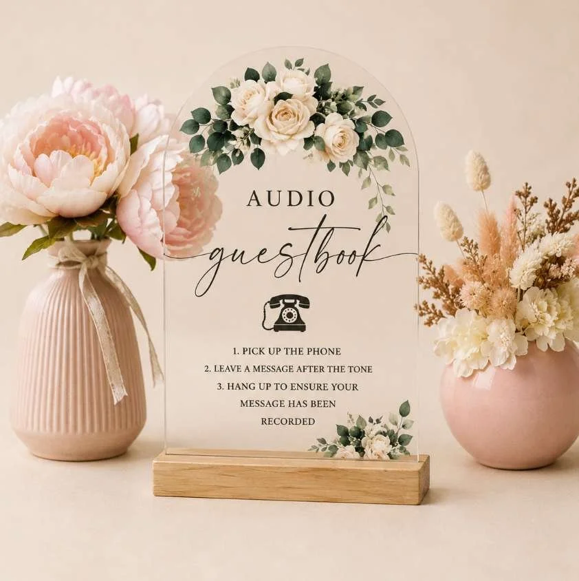 A sign with floral decoration reads 'Audio guestbook' with instructions to pick up the phone, leave a message after the tone, and hang up to ensure the message is recorded, flanked by pink and white flower arrangements.