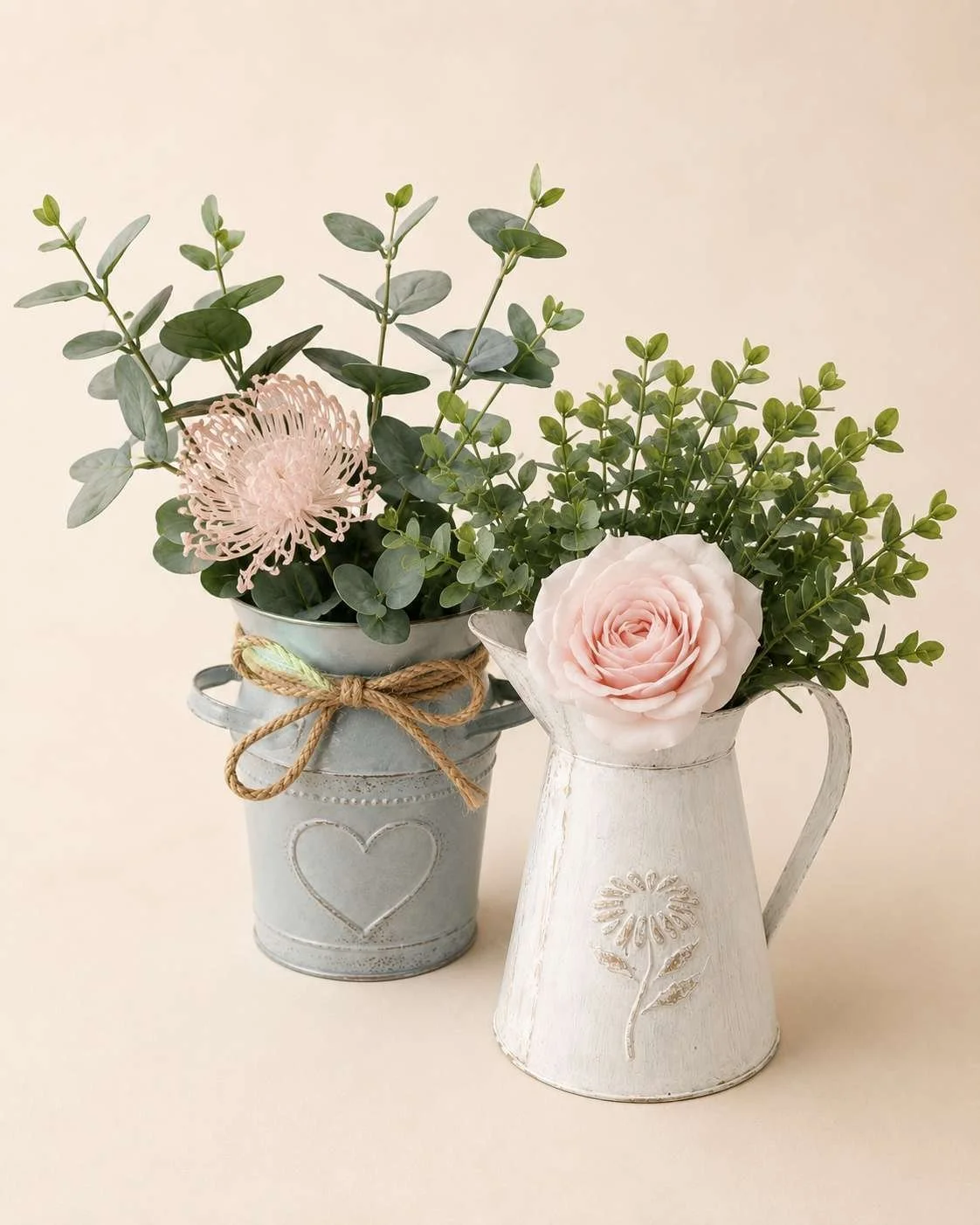 Two vases with pink roses and greenery, one in a gray metal with a heart design and twine bow, the other in a white distressed metal with a floral design, set against a plain beige background.