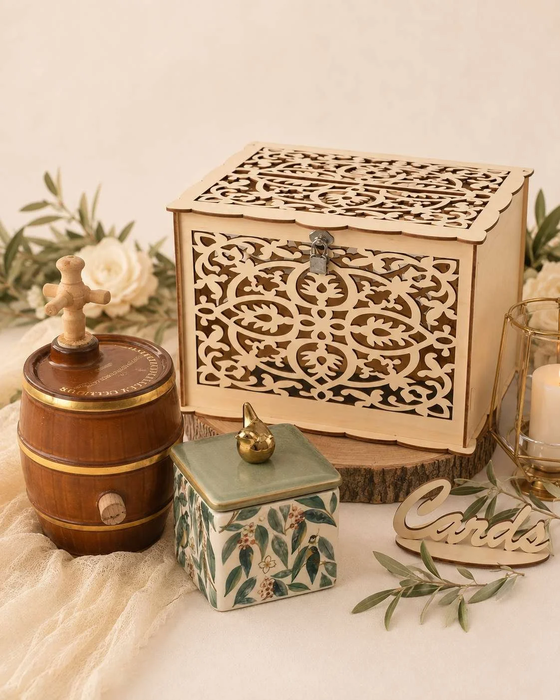Decorative wooden box with intricate laser-cut pattern, a small ceramic container with a floral design and gold lid, a miniature barrel with a wooden top, a gold bird figurine on the ceramic container, a 'Candy' sign, greenery, and candles on a beige tablecloth.