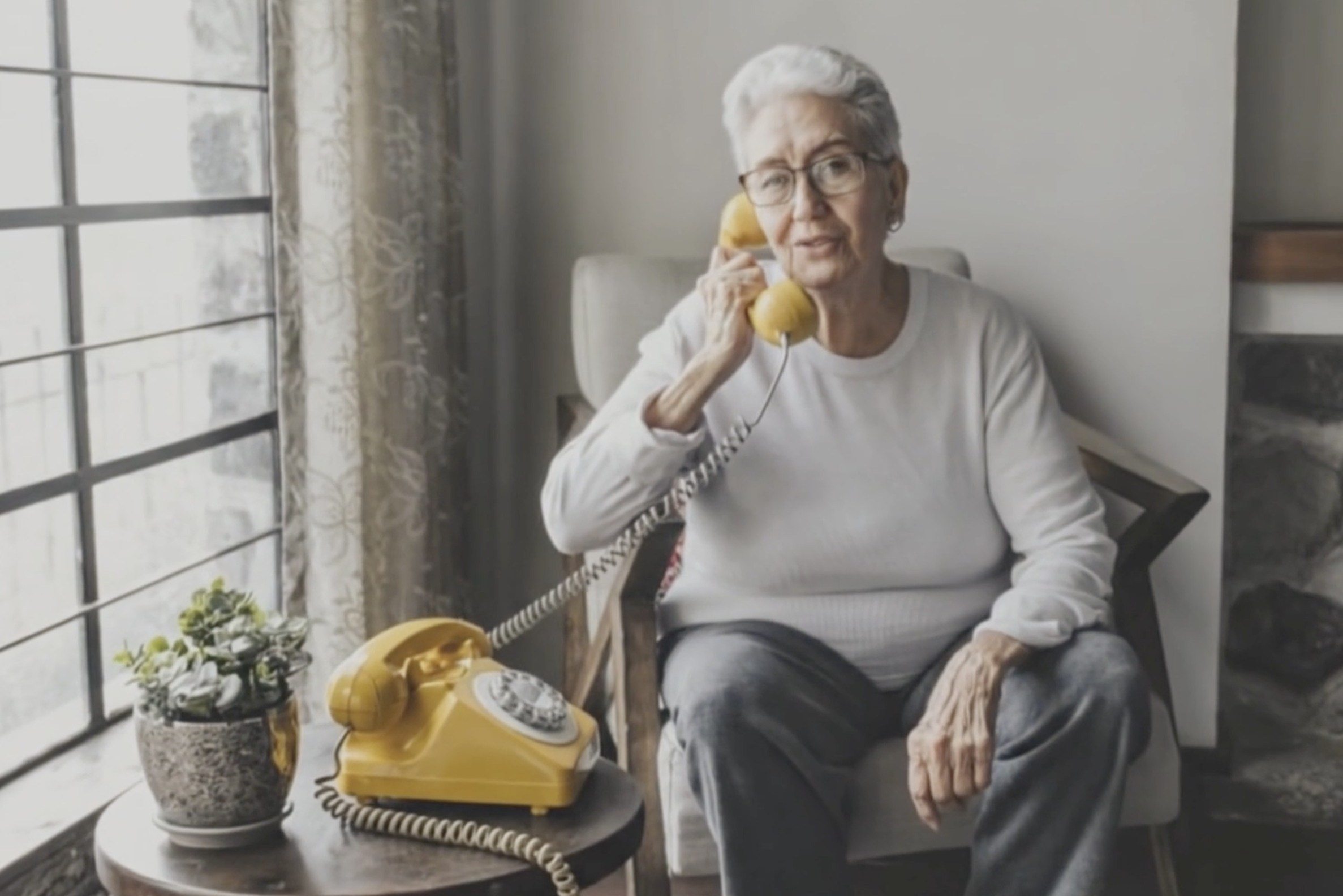 An elderly woman sitting in a beige armchair, smiling while talking on a black rotary phone. There is a wooden side table with a vintage rotary phone, a photo frame, and a vase with flowers in the background. The table in front has framed photographs, a lace doily, a teacup, and a book.