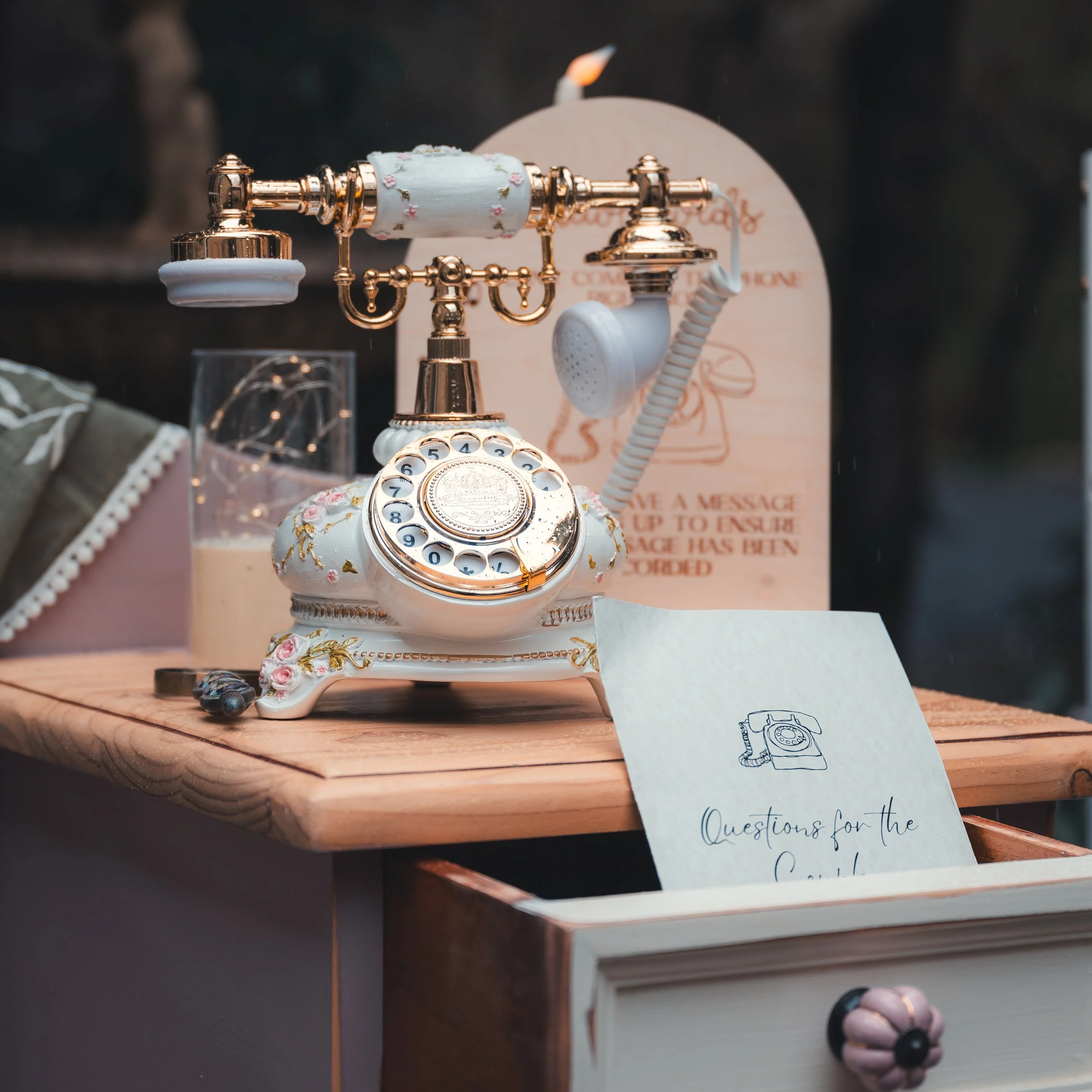 Vintage rotary telephone decorated with pearl accents and floral patterns on a wooden surface, with a handwritten note and a pink decorative item nearby.