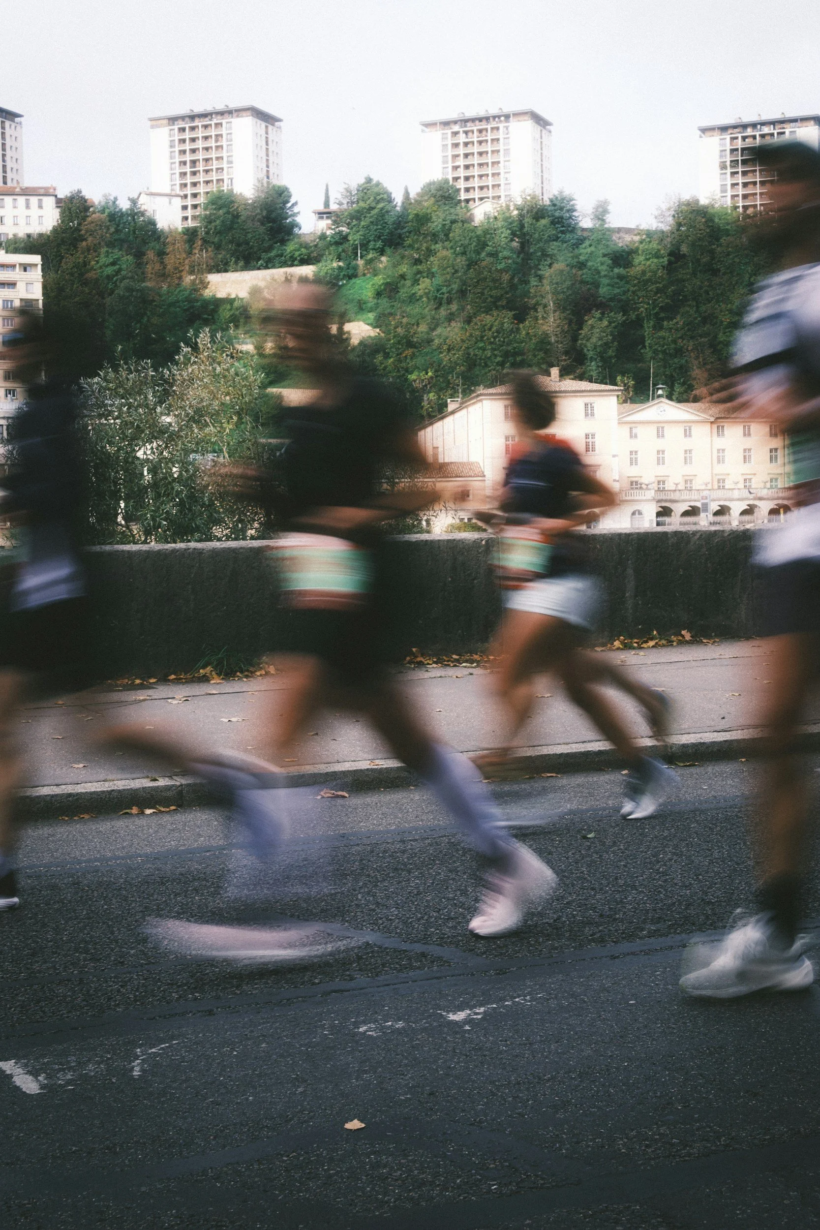 Blurred motion of four runners in athletic gear running on a city street with a background of trees and tall residential buildings.