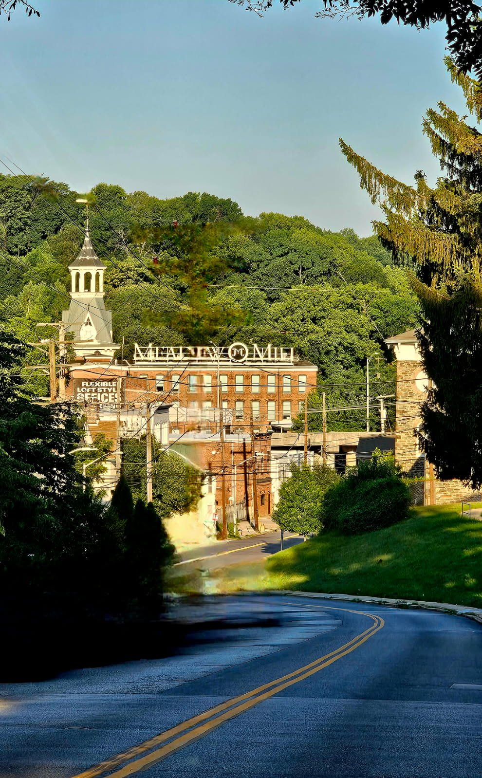 A winding road leading into a small town with a brick building labeled 'Meadow Mill' in the background. The building has a sign advertising flexible loft styles and offices. A church steeple is visible on the left, surrounded by green trees, with electric wires crossing the scene.