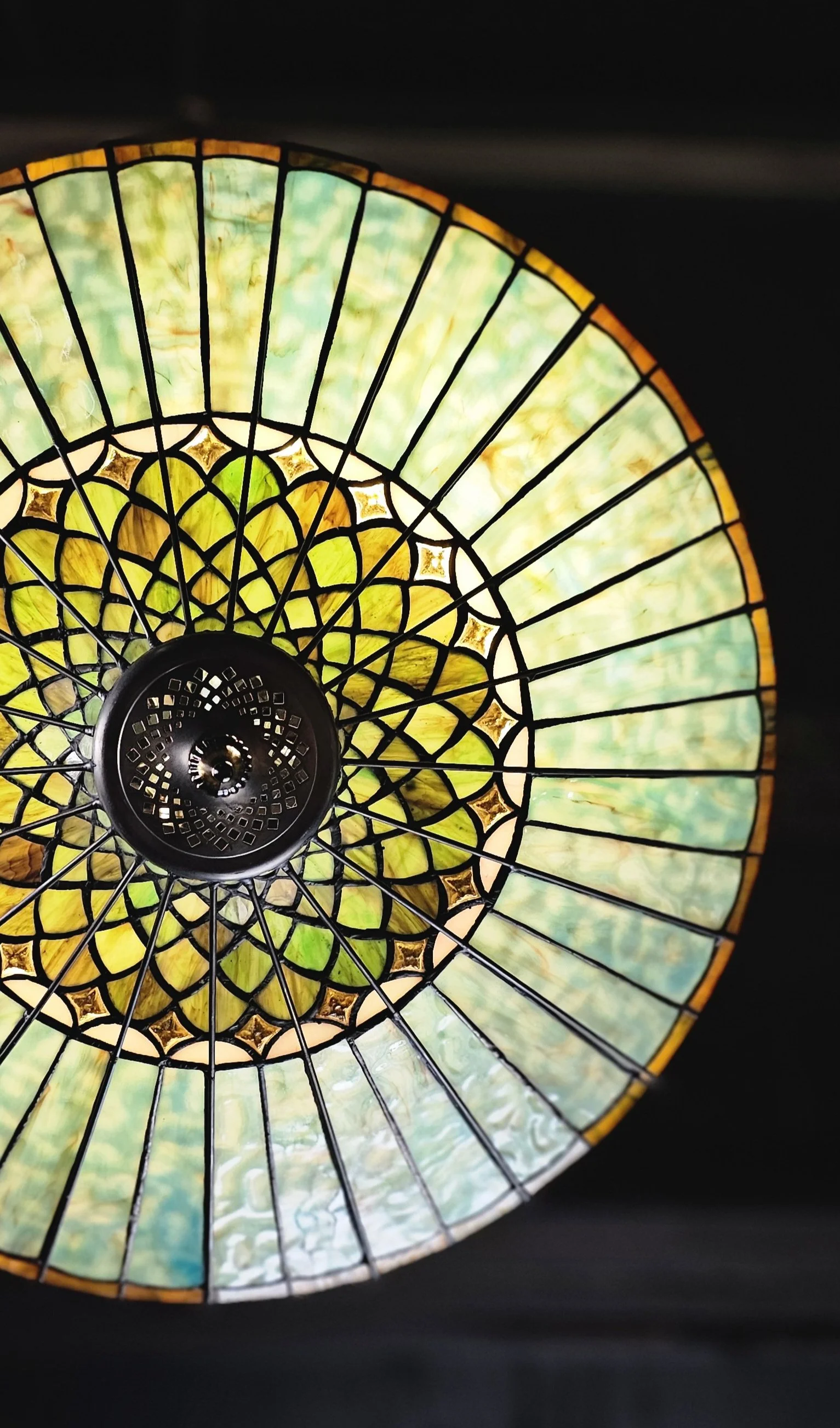 Close-up of a stained glass ceiling light fixture with yellow, green, and blue glass panels, supported by black metal framing, against a dark background.