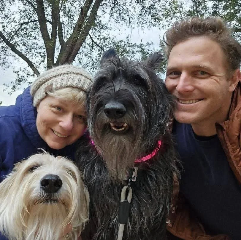A family of two adults and two dogs taking a selfie outdoors with trees in the background.